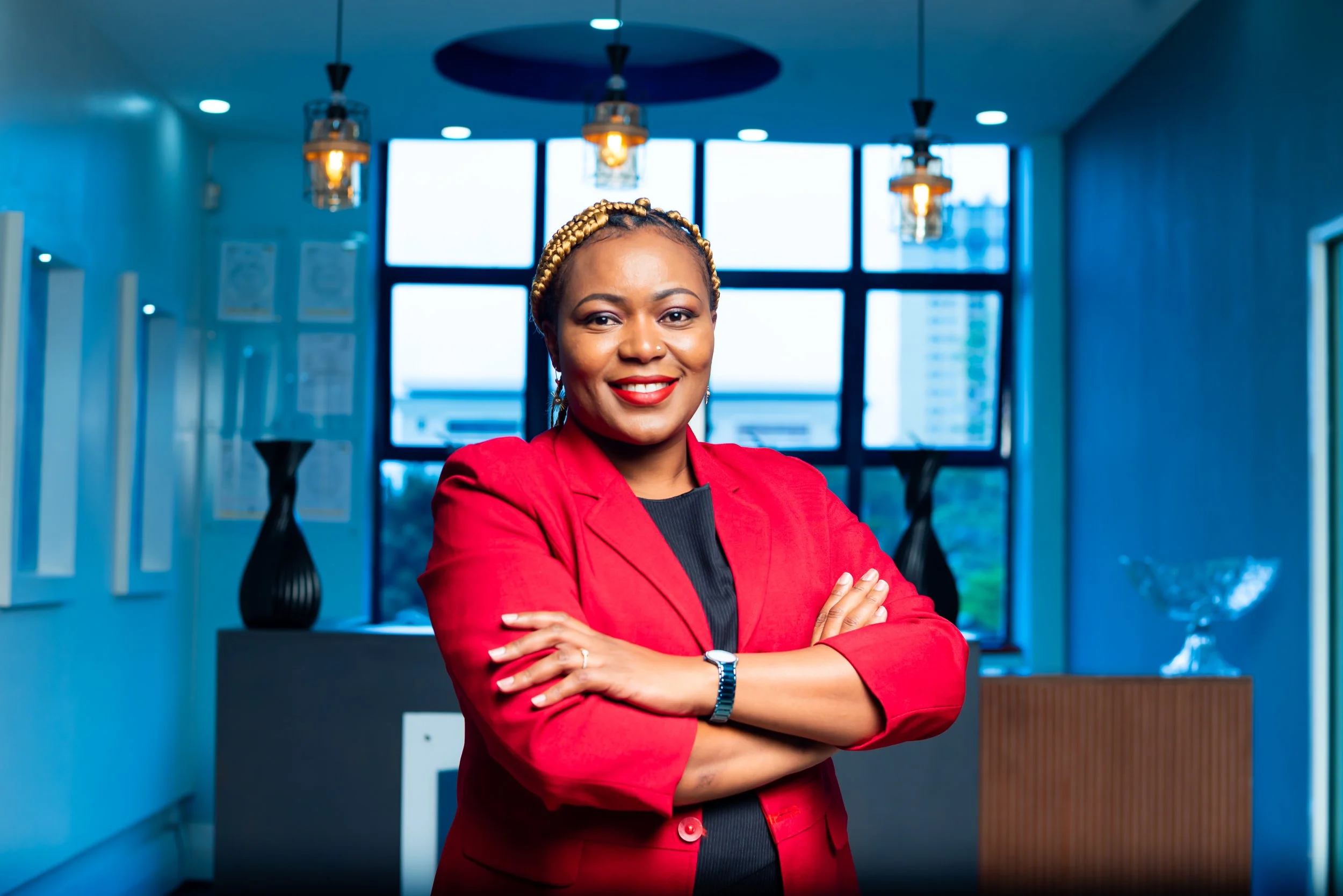 A confident woman in a red blazer standing with arms crossed in a modern office space with large windows and blue walls.