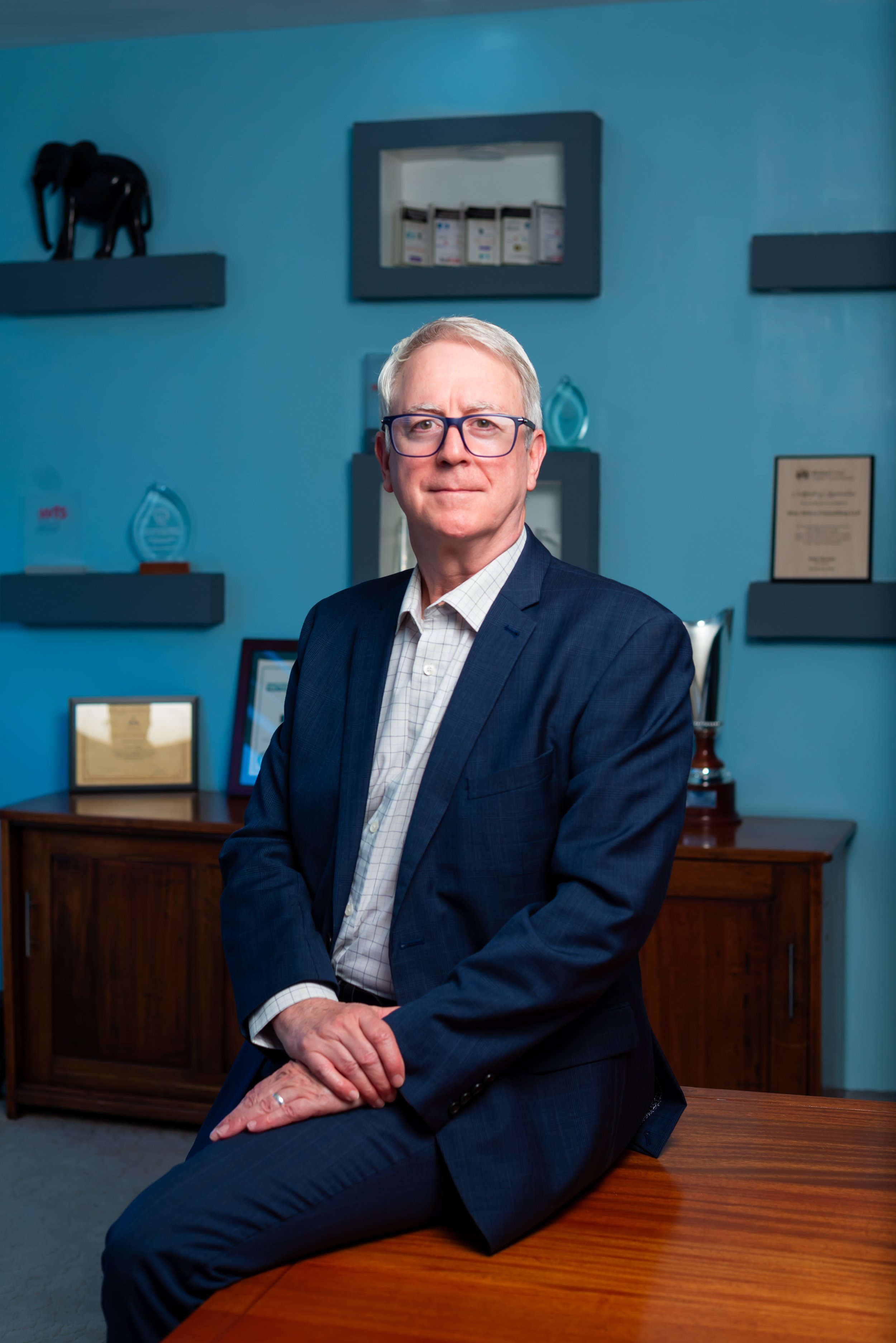 Mick Murphy, Viva's Senior Partner sitting on a wooden table in an office with blue walls. The background features framed certificates, awards, and shelves with trophies and decorative items.