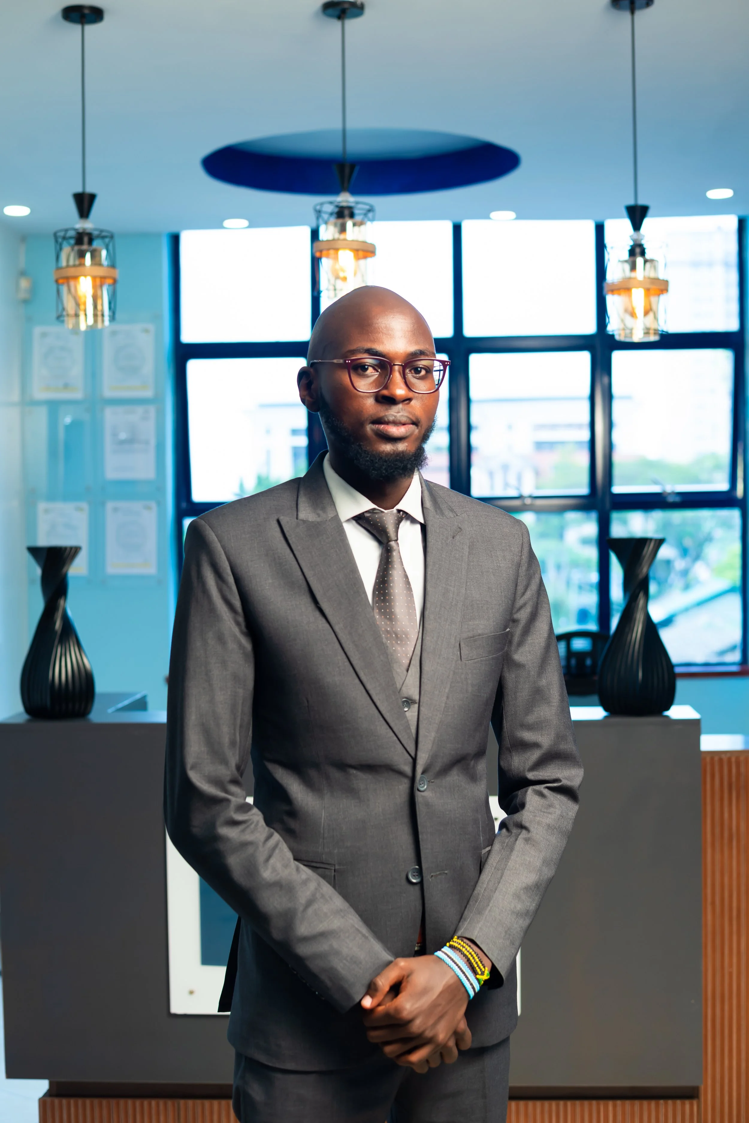 A man in a gray suit with glasses standing in an office lobby or reception area with large windows and modern lighting fixtures.