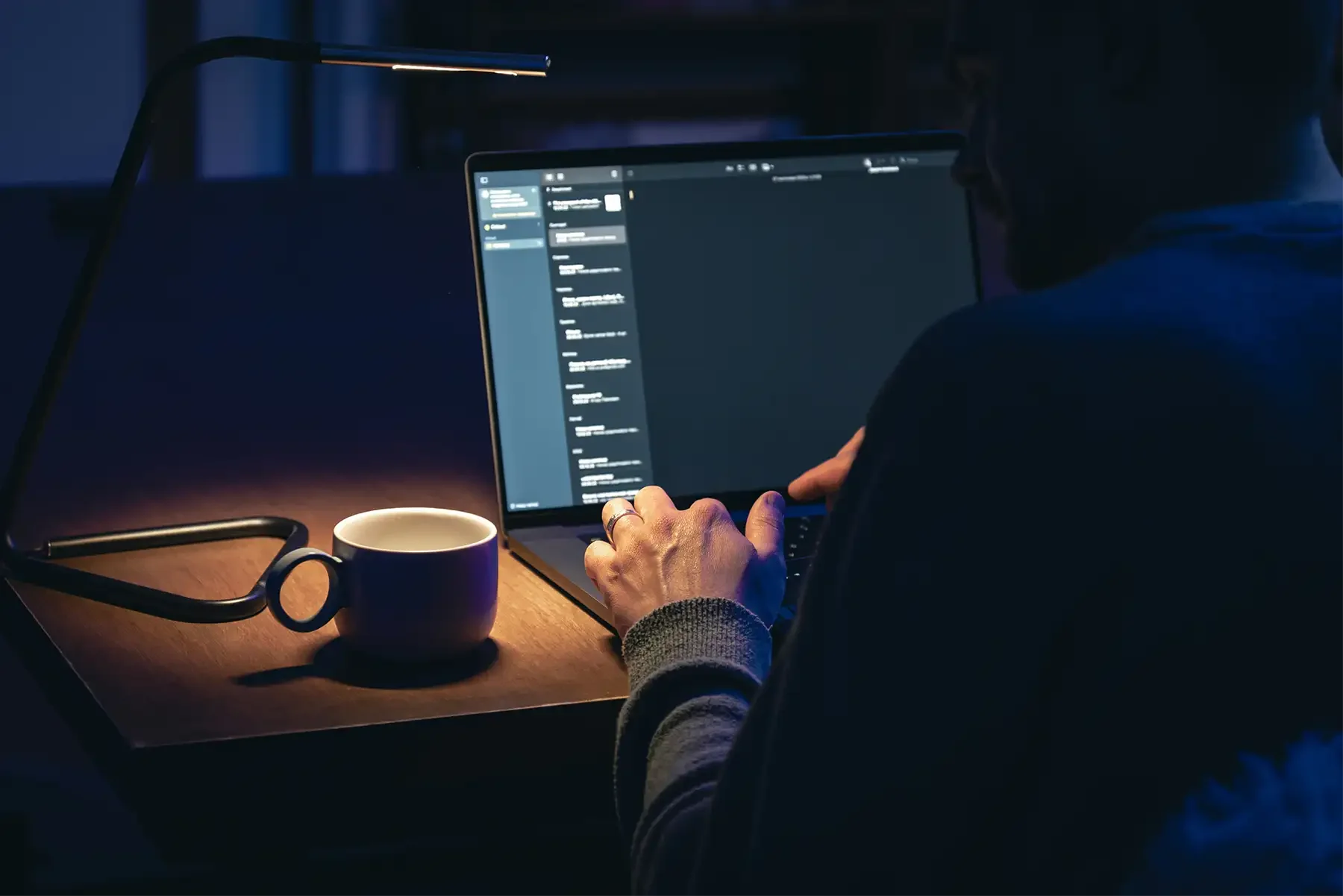 A person working on a laptop at a desk in a dimly lit room, with a desk lamp and a coffee mug nearby.