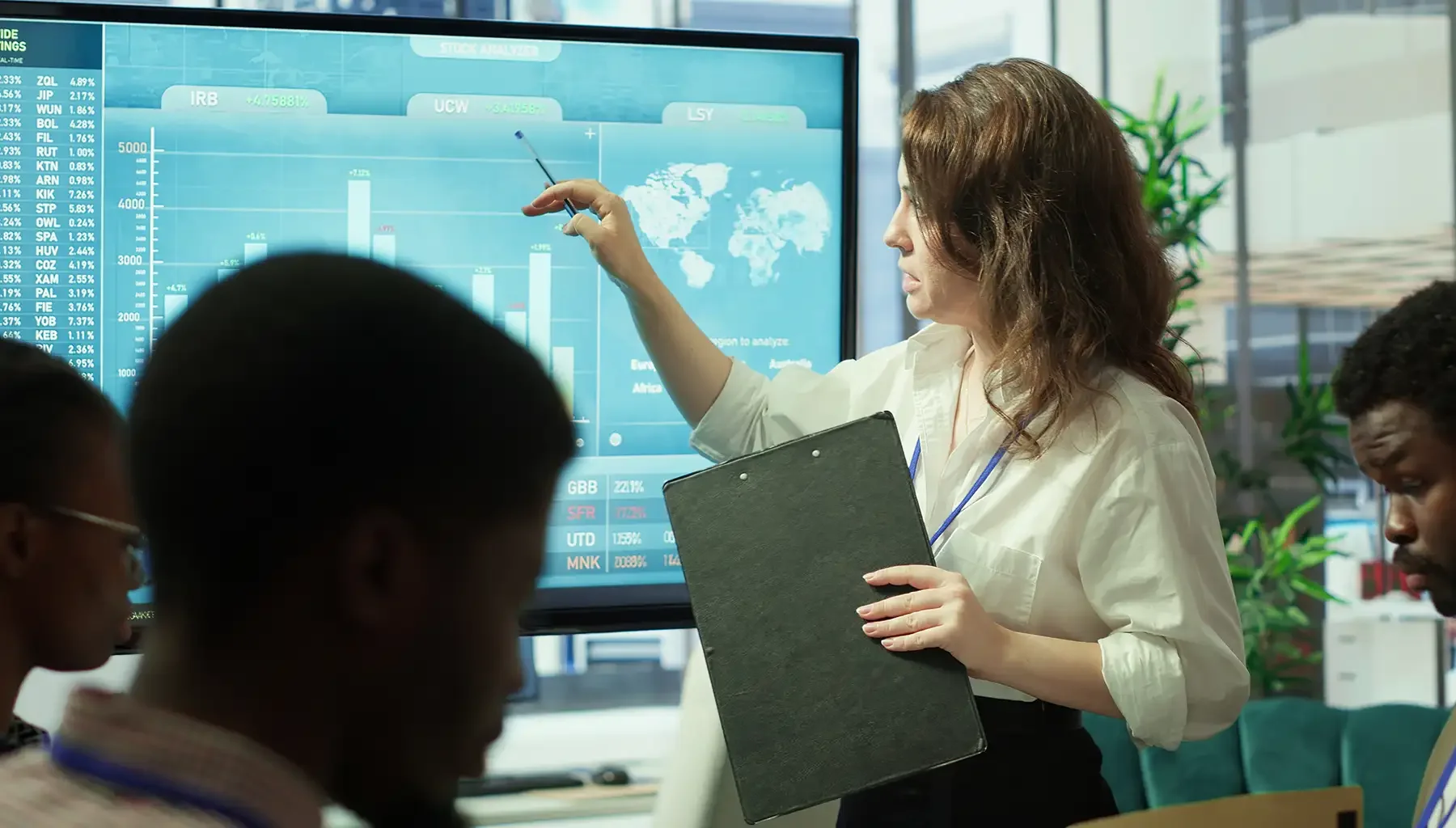 A woman presenting financial data and stock market graphs on a large screen to a group of people in a conference room.