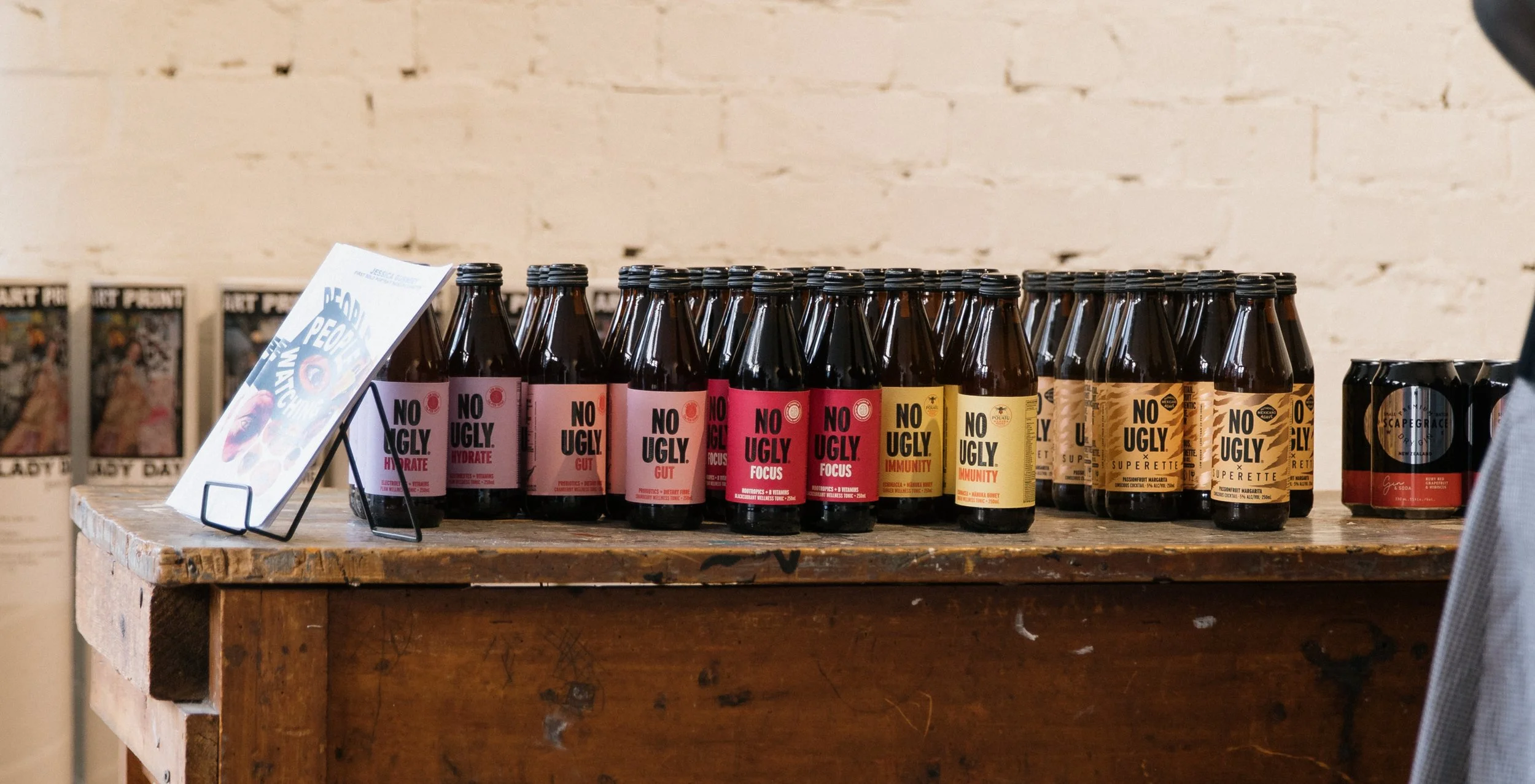 A display of bottled beverages with labels reading 'No Ugly' and additional descriptive words, arranged on a rustic wooden table with a promotional card stand.