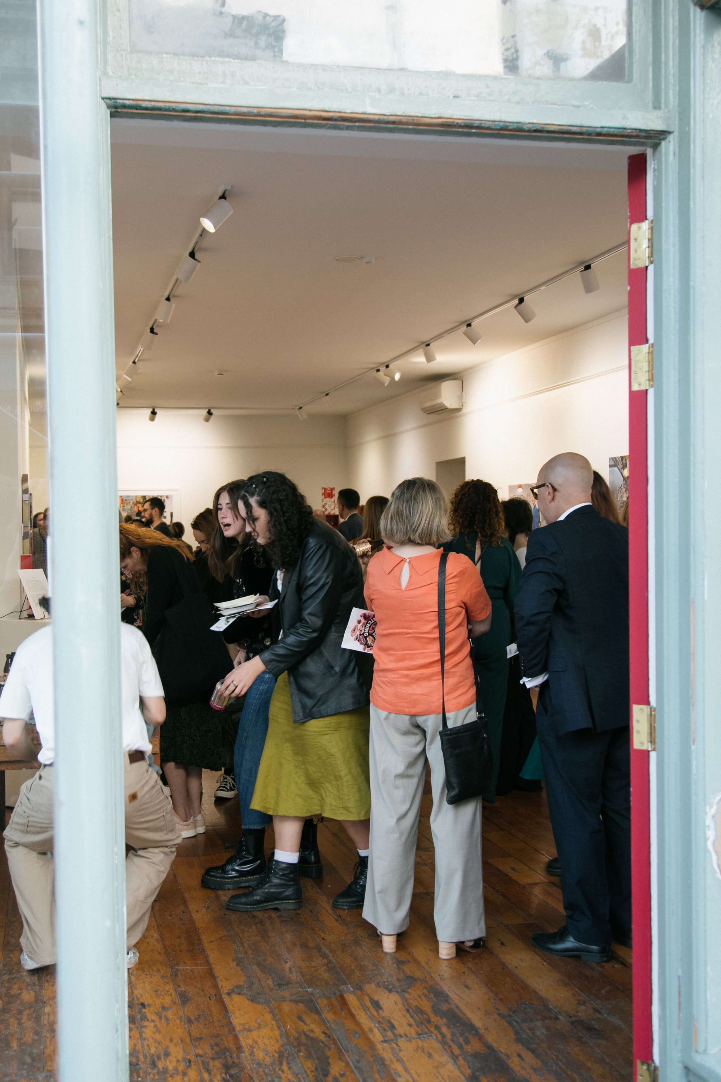 People attending an art gallery or exhibition, viewing artwork displayed on white walls, with diverse individuals engaging and conversing inside a well-lit room.