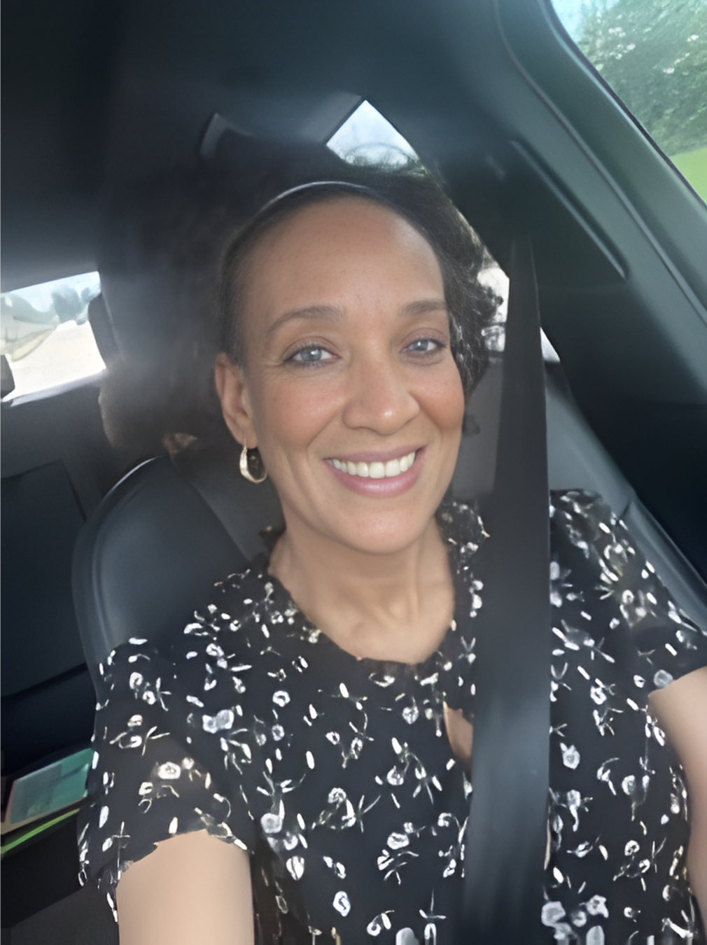 Smiling woman in a patterned dress sitting in a car, taking a selfie.