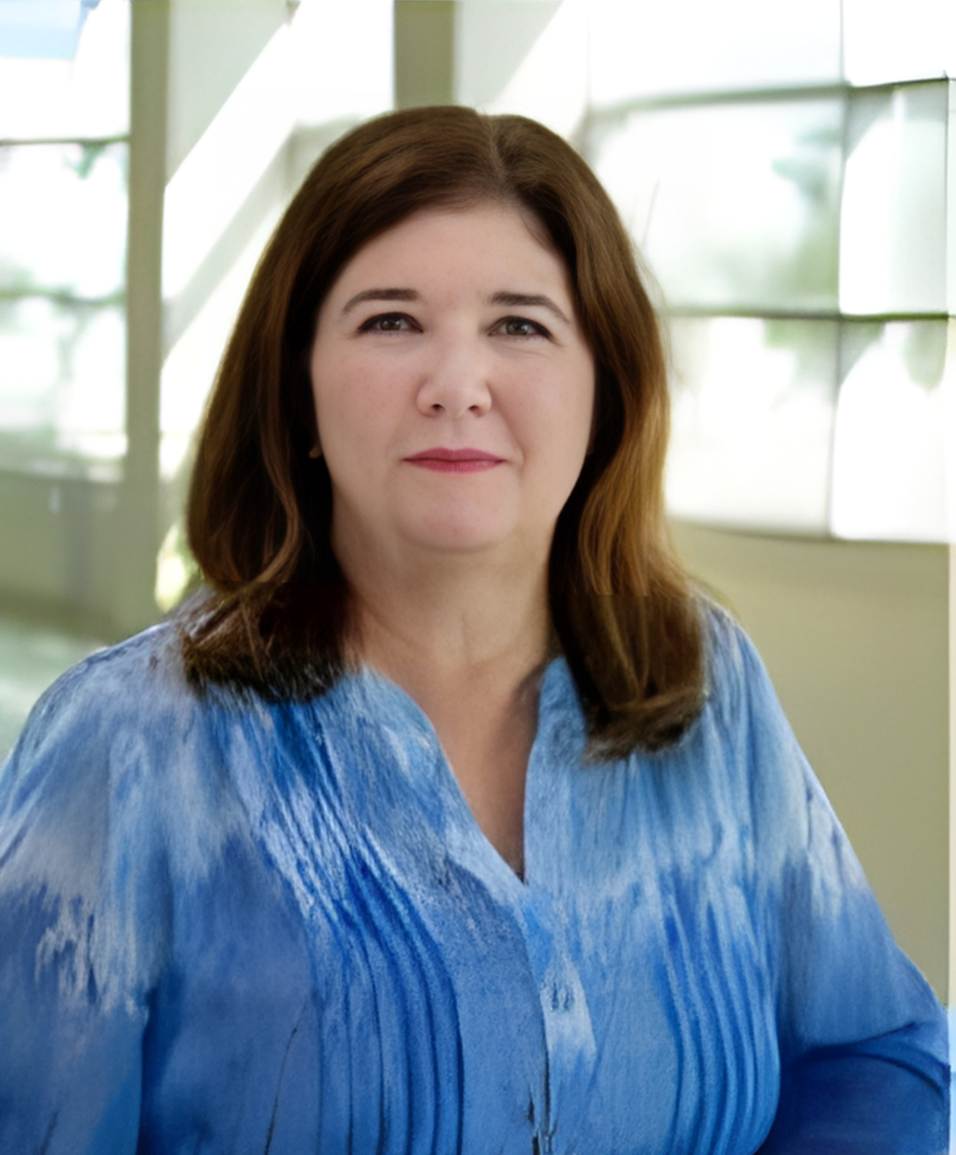 A woman with medium-length brown hair and fair skin, wearing a blue blouse, standing indoors near a window with blinds.
