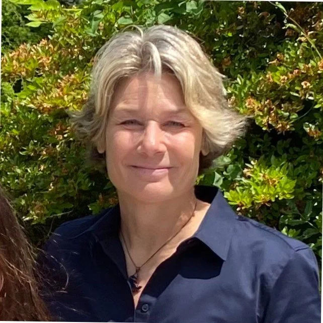 A woman with short blonde hair smiling outdoors, standing in front of green and brown bush foliage.