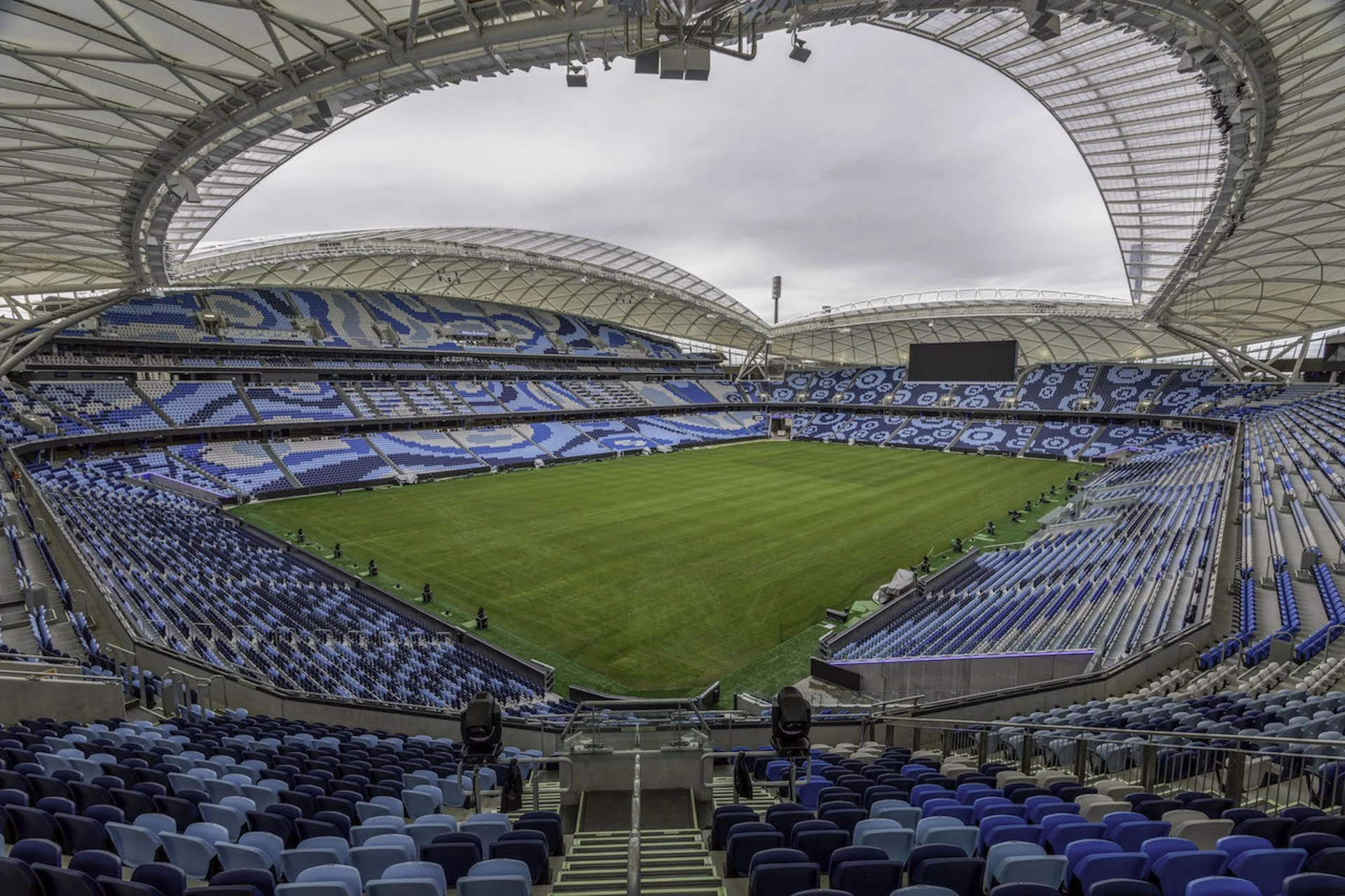 Empty soccer stadium with blue seats, green field, and cloudy sky overhead.