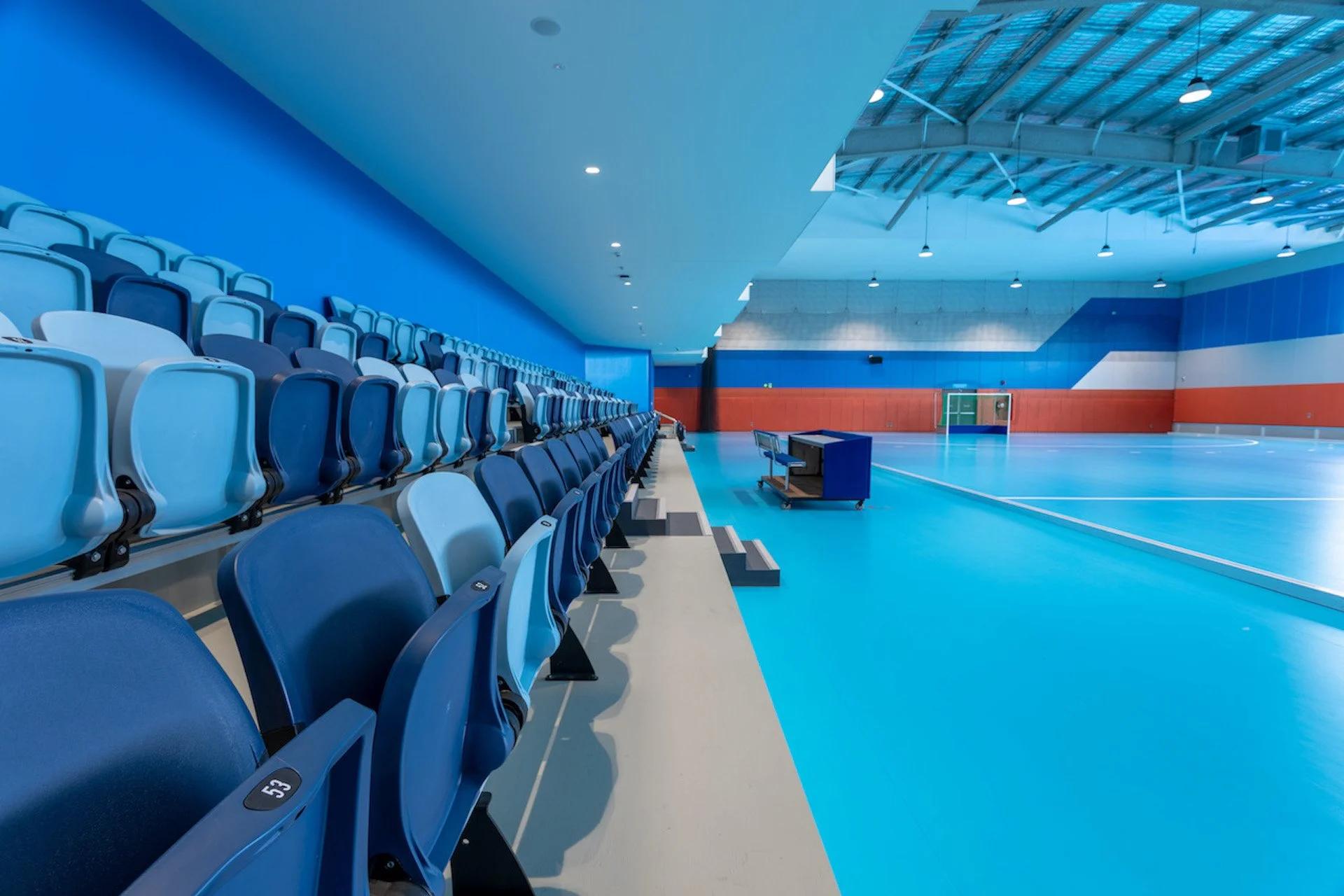 Empty indoor sports arena with blue and white seating along the side, a polished blue floor with court markings, and high ceiling with metal beams and hanging lights.