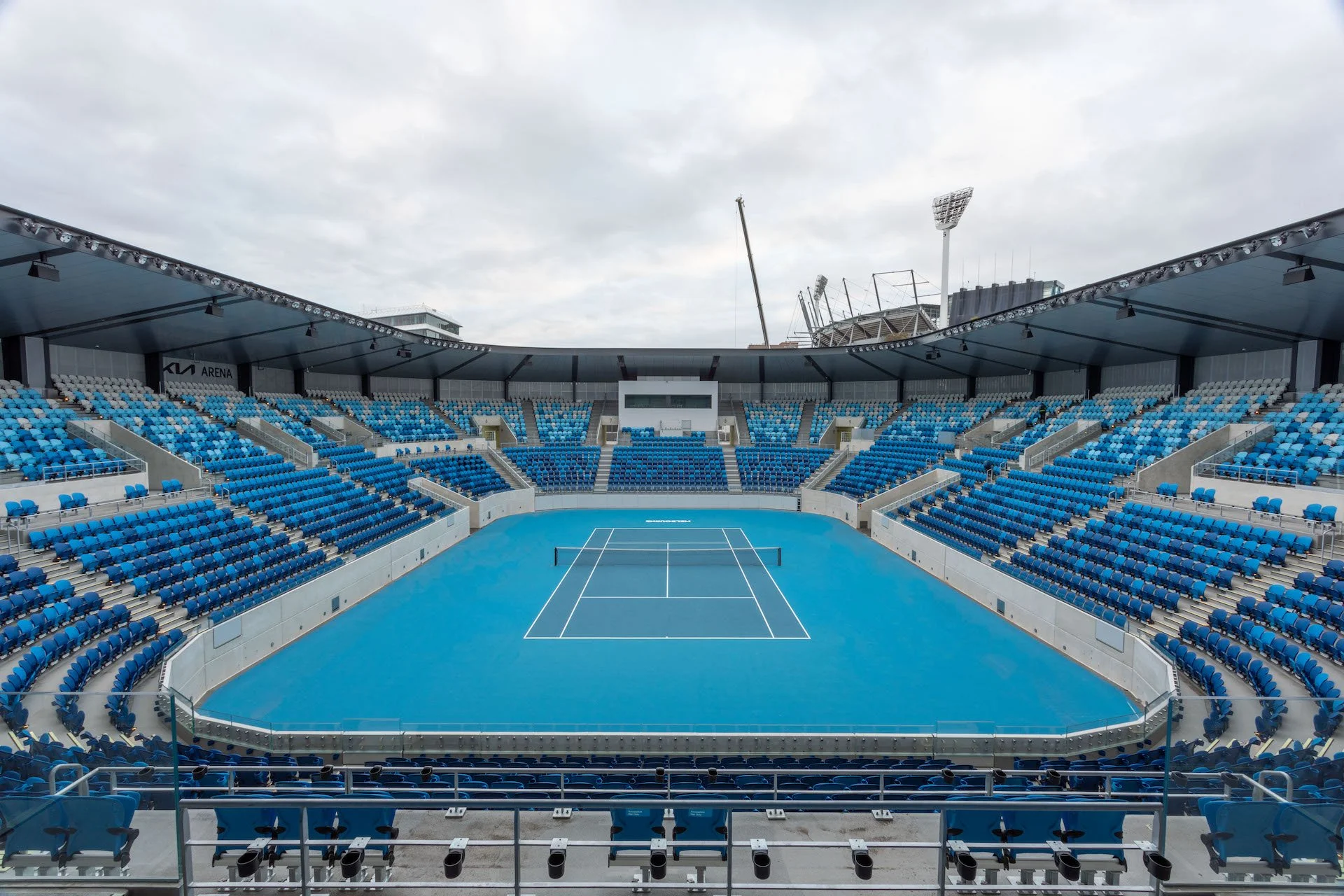 Empty blue tennis court in a stadium with blue seats, covered roof, and overcast sky.