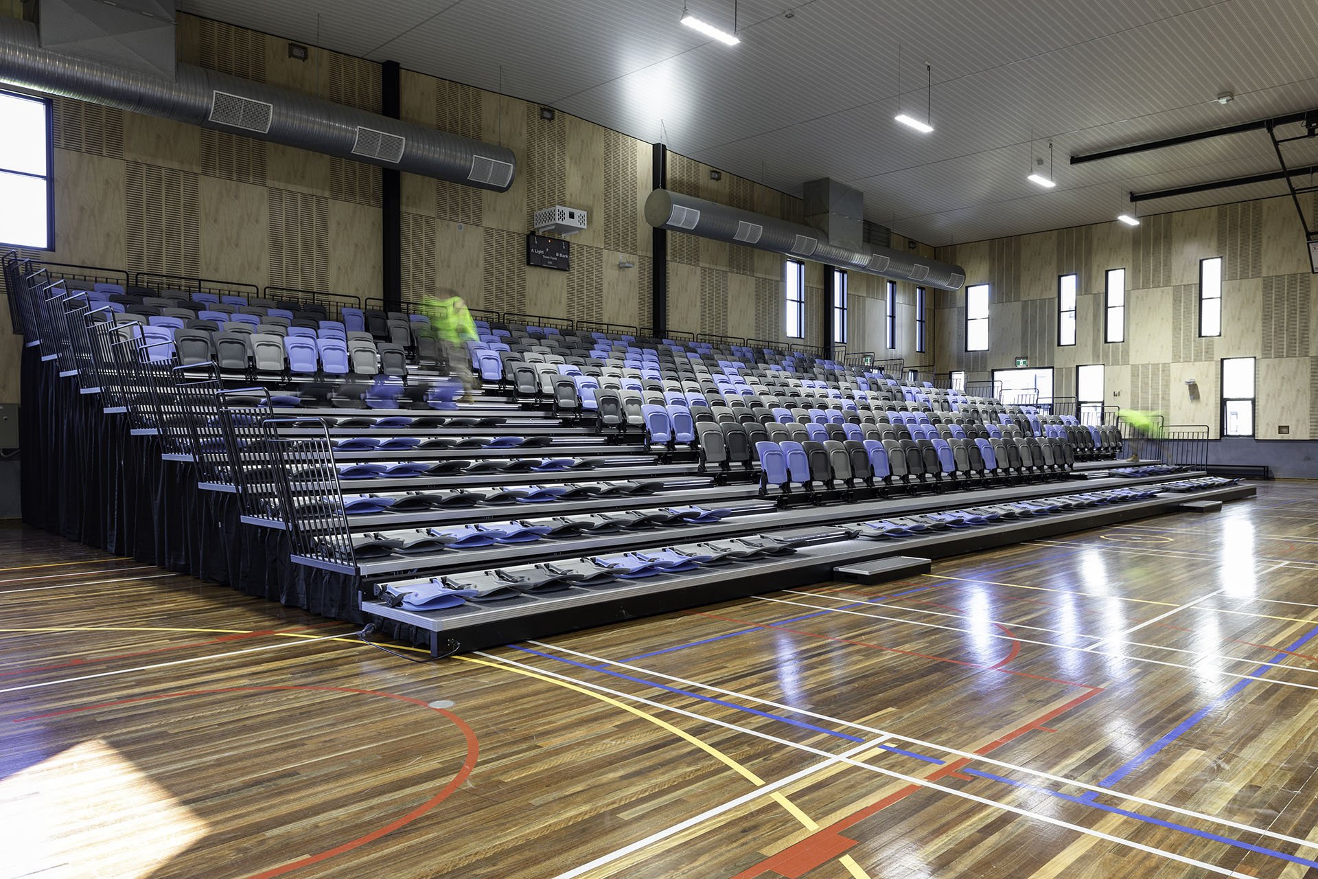 Empty indoor gymnasium with tiered seating and wooden flooring, with a few blurred figures moving on the bleachers.