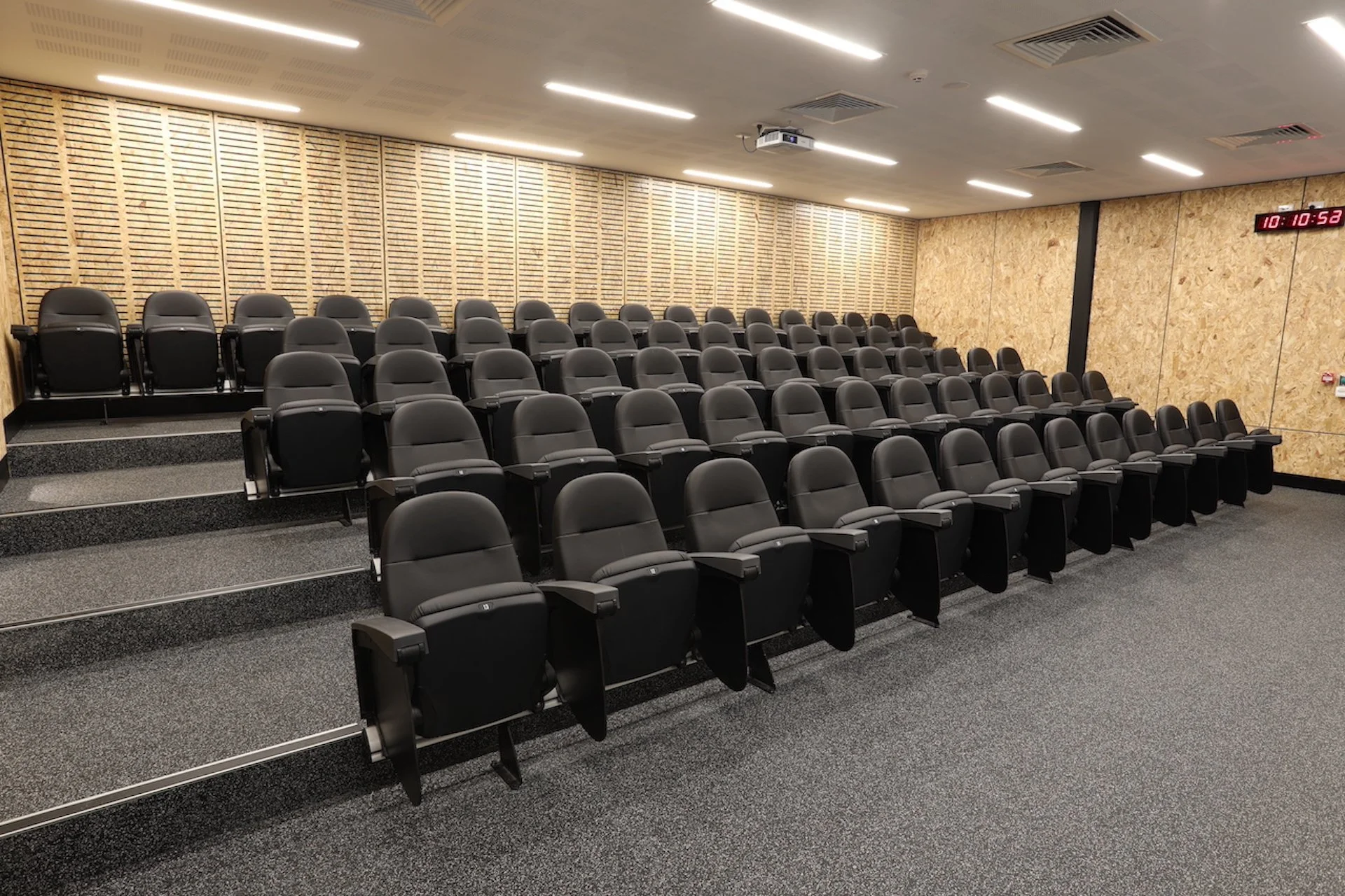 Empty lecture hall with black cushioned seats arranged in ascending rows, gray carpeted floor, wooden wall paneling on one side, and a digital clock showing 10:10.