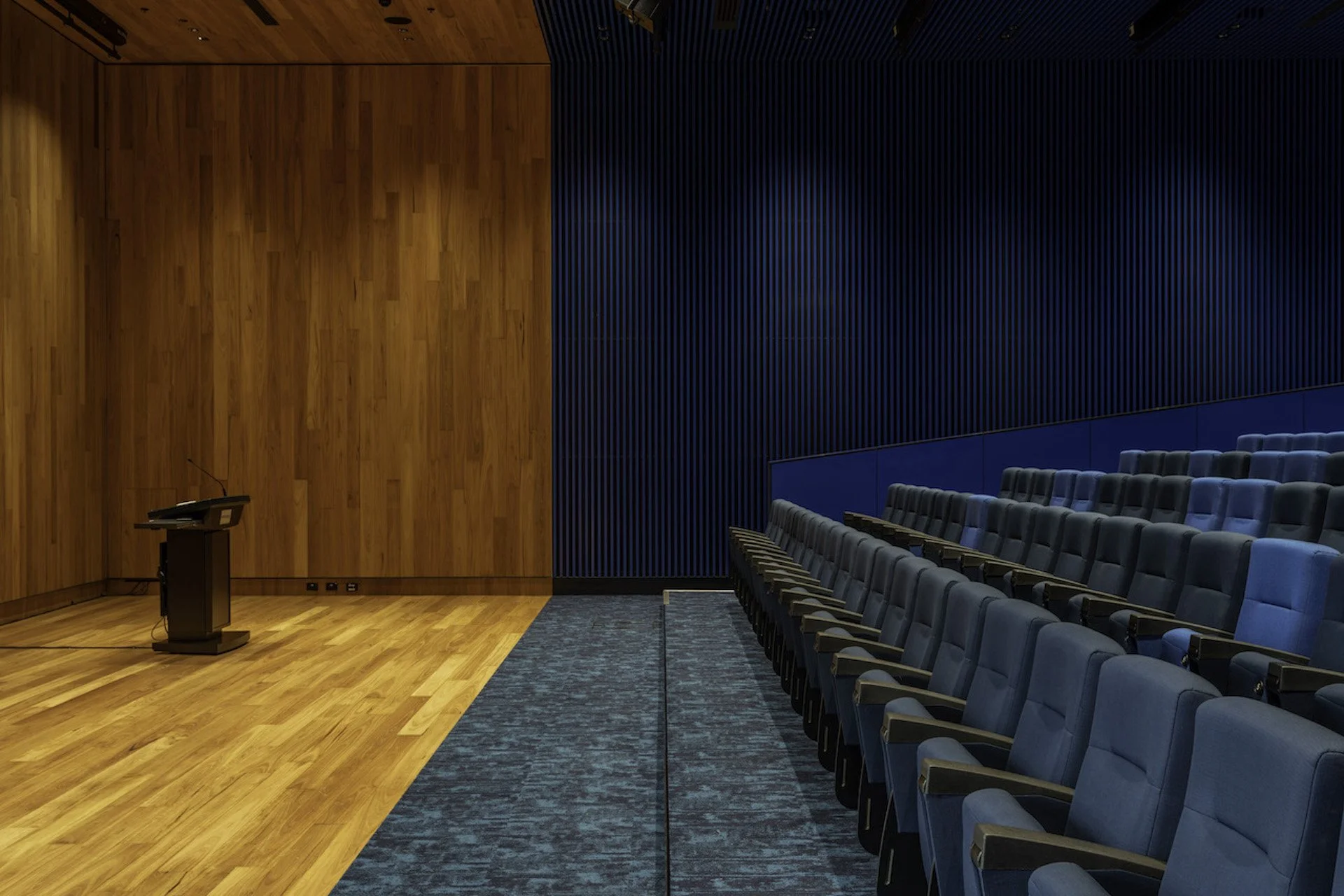 An empty auditorium with a wooden stage, a podium, blue upholstered seats, and wooden wall paneling.