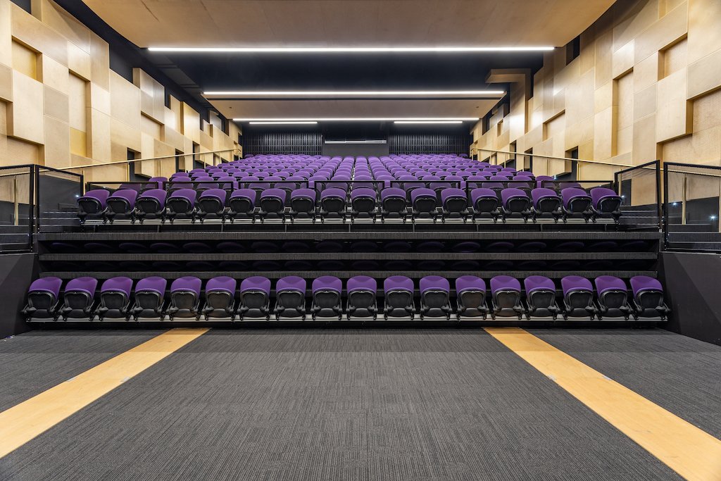 An empty auditorium with rows of purple seats, black railings, and a patterned beige and black wall.