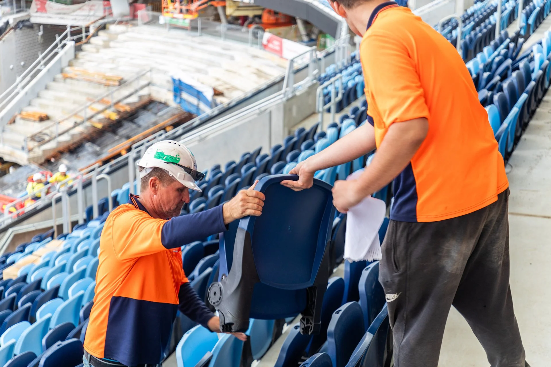 Two workers in orange and blue uniforms installing or repairing blue stadium seats in a sports arena.
