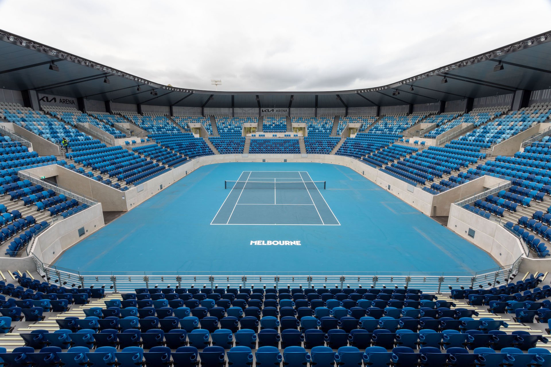 Empty tennis stadium with blue court and seating, marking 'Melbourne' on court, under an overcast sky.
