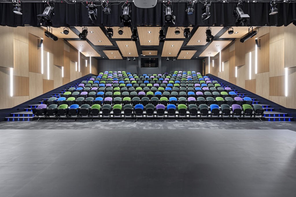 Empty theater with rows of colorful chairs on a stage, black ceiling with lights, wood-paneled walls, and steps on both sides.