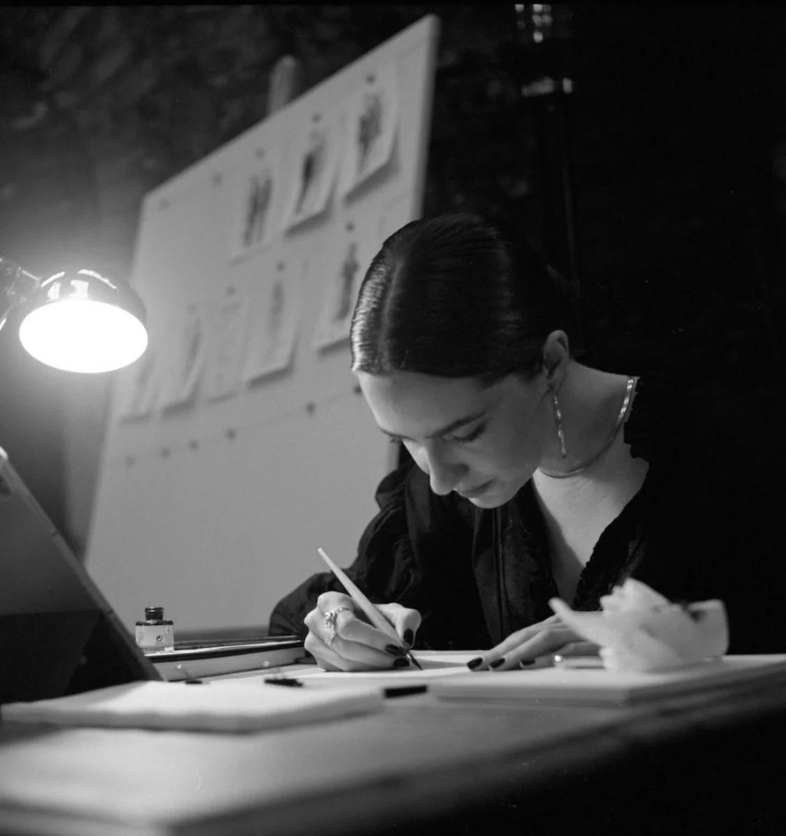 A woman with dark, slicked-back hair writing at a desk under a bright desk lamp, with a blurred bulletin board in the background.