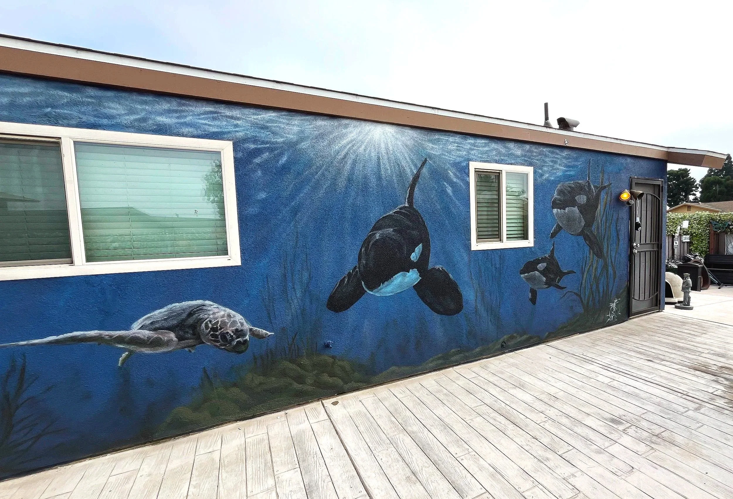Colorful mural of underwater scene with three orcas swimming in front of a blue background, on the side of a building with two windows and a door, in an outdoor area with a wooden deck.
