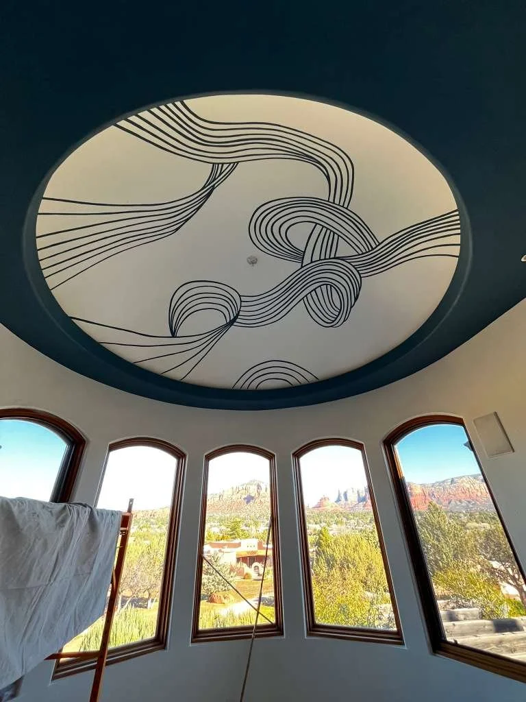 Ceiling with a modern black and white line art design, viewed from a room with large arched windows looking out at a scenic landscape with red rock formations, trees, and a clear blue sky.