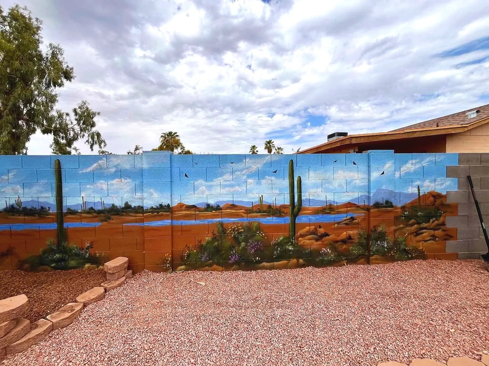 A residential desert mural by Jade Bamford Murals in Tempe AZ, featuring a painted block wall with a Sonoran desert landscape, saguaro cacti, blooming wildflowers, a winding path, and distant mountains under a blue sky.