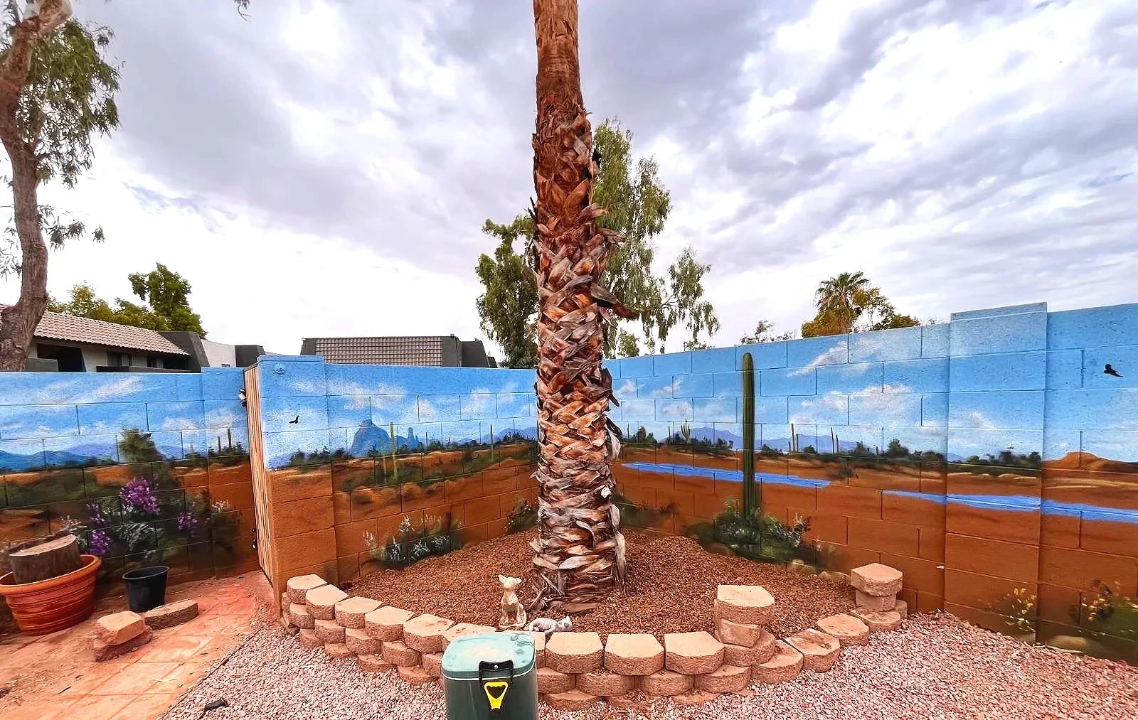 A residential desert mural by Jade Bamford Murals in Tempe AZ, featuring a painted block wall with a Sonoran desert landscape, saguaro cacti, blooming wildflowers, a winding path, and distant mountains under a blue sky.