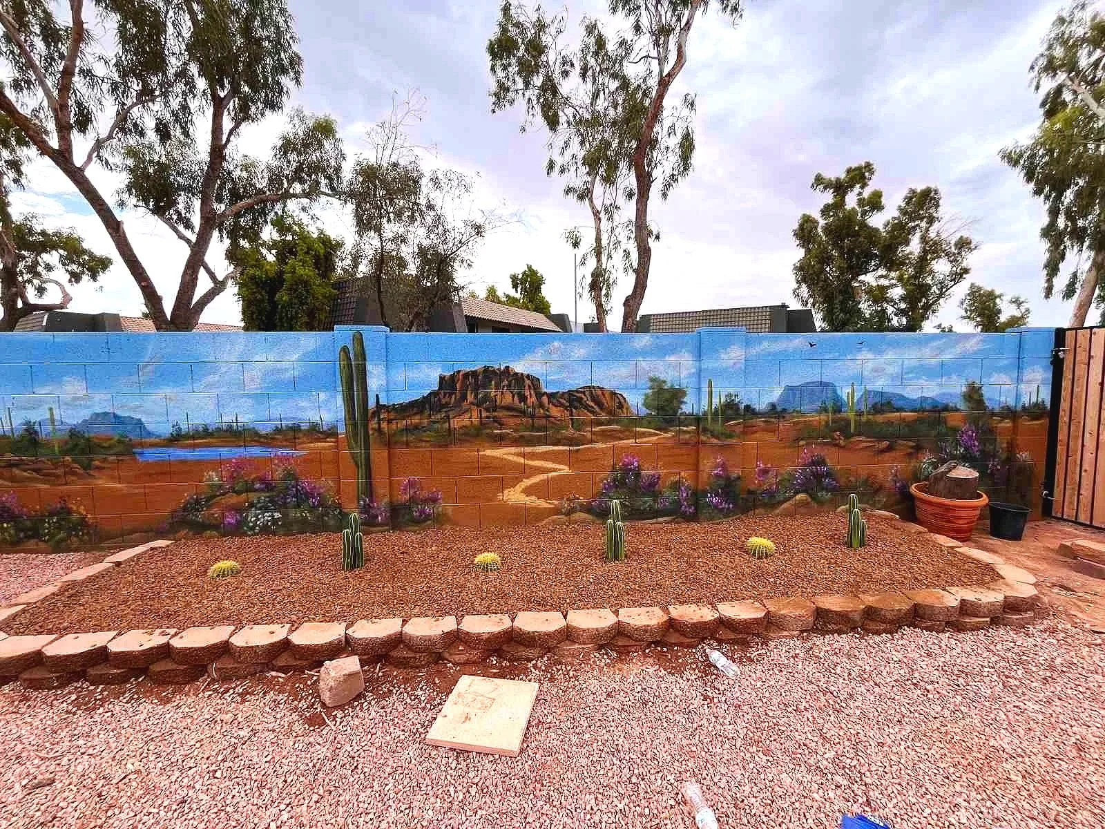 A residential desert mural by Jade Bamford Murals in Tempe AZ, featuring a painted block wall with a Sonoran desert landscape, saguaro cacti, blooming wildflowers, a winding path, and distant mountains under a blue sky.