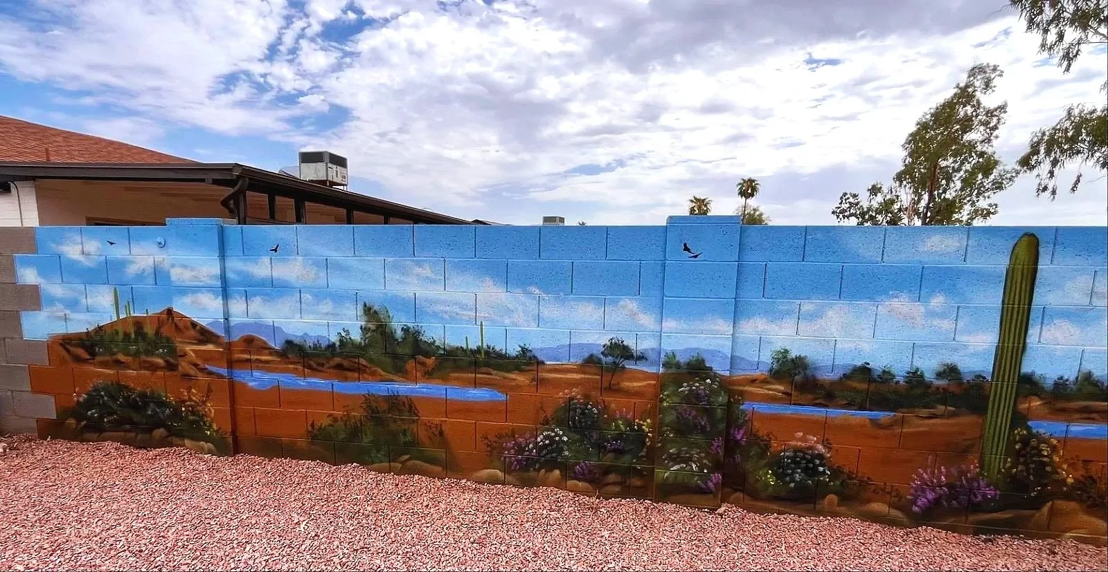 A residential desert mural by Jade Bamford Murals in Tempe AZ, featuring a painted block wall with a Sonoran desert landscape, saguaro cacti, blooming wildflowers, a winding path, and distant mountains under a blue sky.
