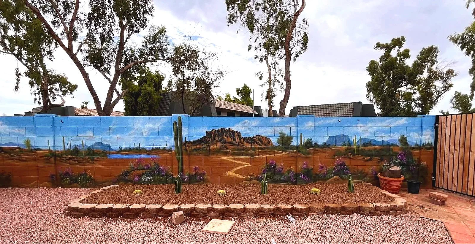 A residential desert mural by Jade Bamford Murals in Tempe AZ, featuring a painted block wall with a Sonoran desert landscape, saguaro cacti, blooming wildflowers, a winding path, and distant mountains under a blue sky.