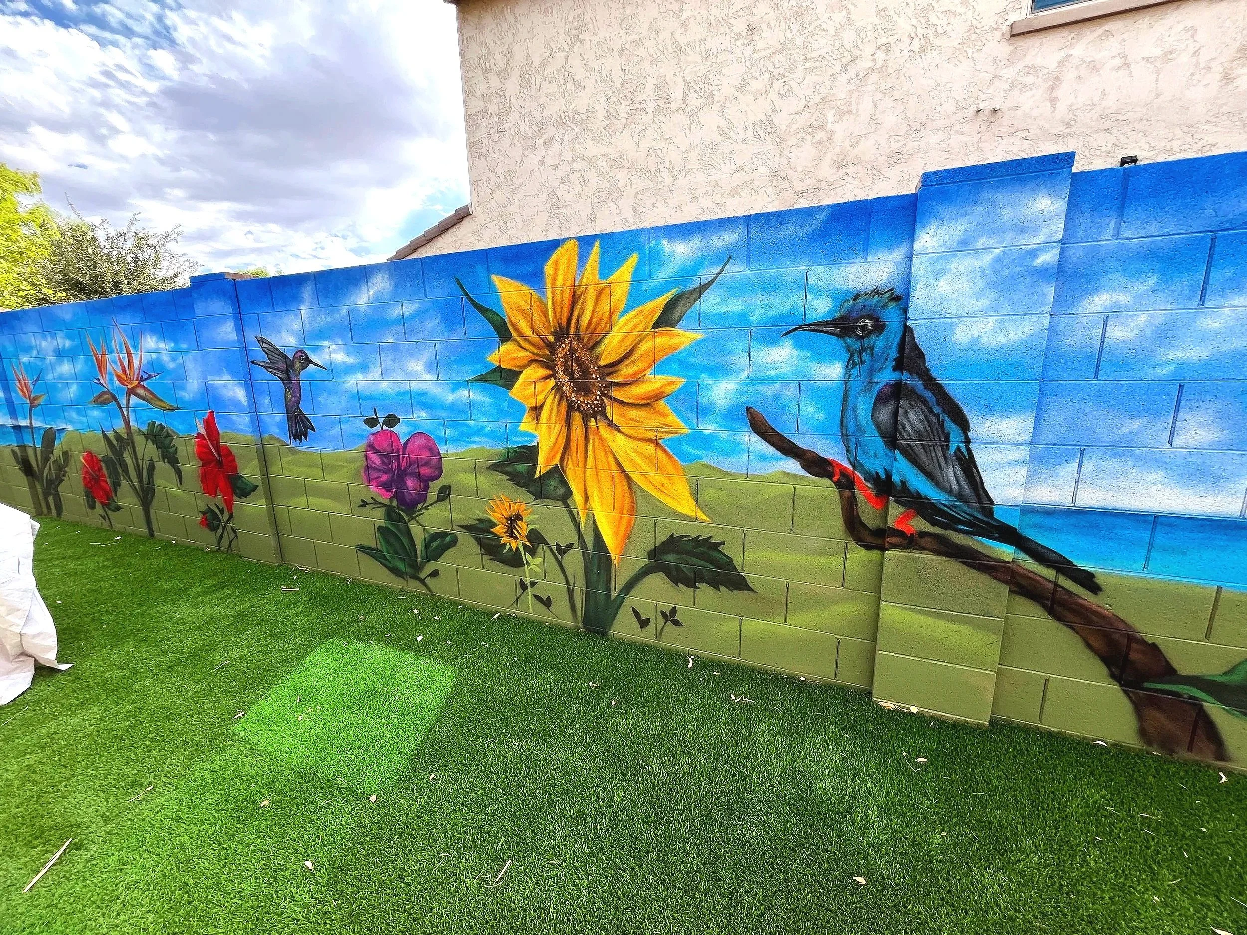 A residential floral mural by Jade Bamford Murals in Surprise AZ, featuring a sunflower, hummingbird, blue bird, and a bright sky.