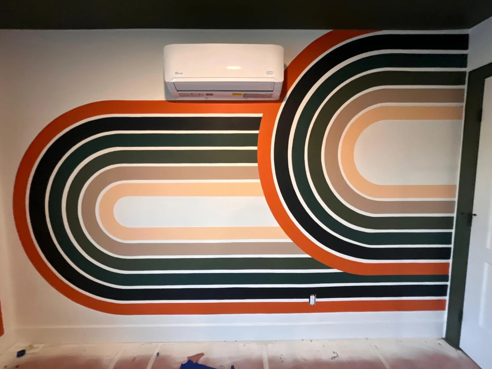 A wall mural with curved, concentric lines in black, white, beige, and orange, resembling an abstract retro design. An air conditioning unit is mounted at the top of the wall.