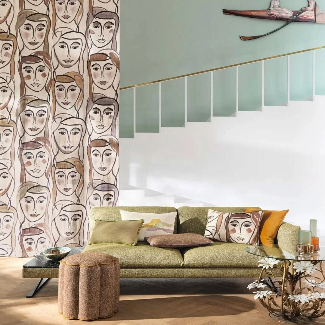 Interno di un soggiorno con pareti decorate, divano verde con cuscini e tavolini, decorazioni floreali, e una scalinata sul soffitto.