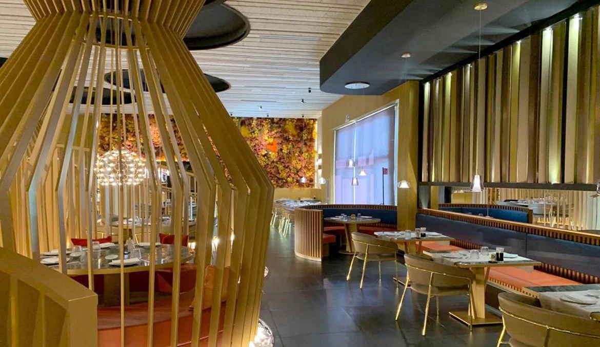 Interno di un ristorante elegante con arredi moderni, sedie dorate, soffitto in legno e decorazioni floreali alle pareti.