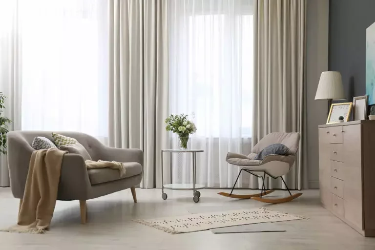 Soggiorno con divano beige, sedia a dondolo, tavolino con vaso di fiori, credenza con lampada, quadri alle pareti e tende leggere bianco