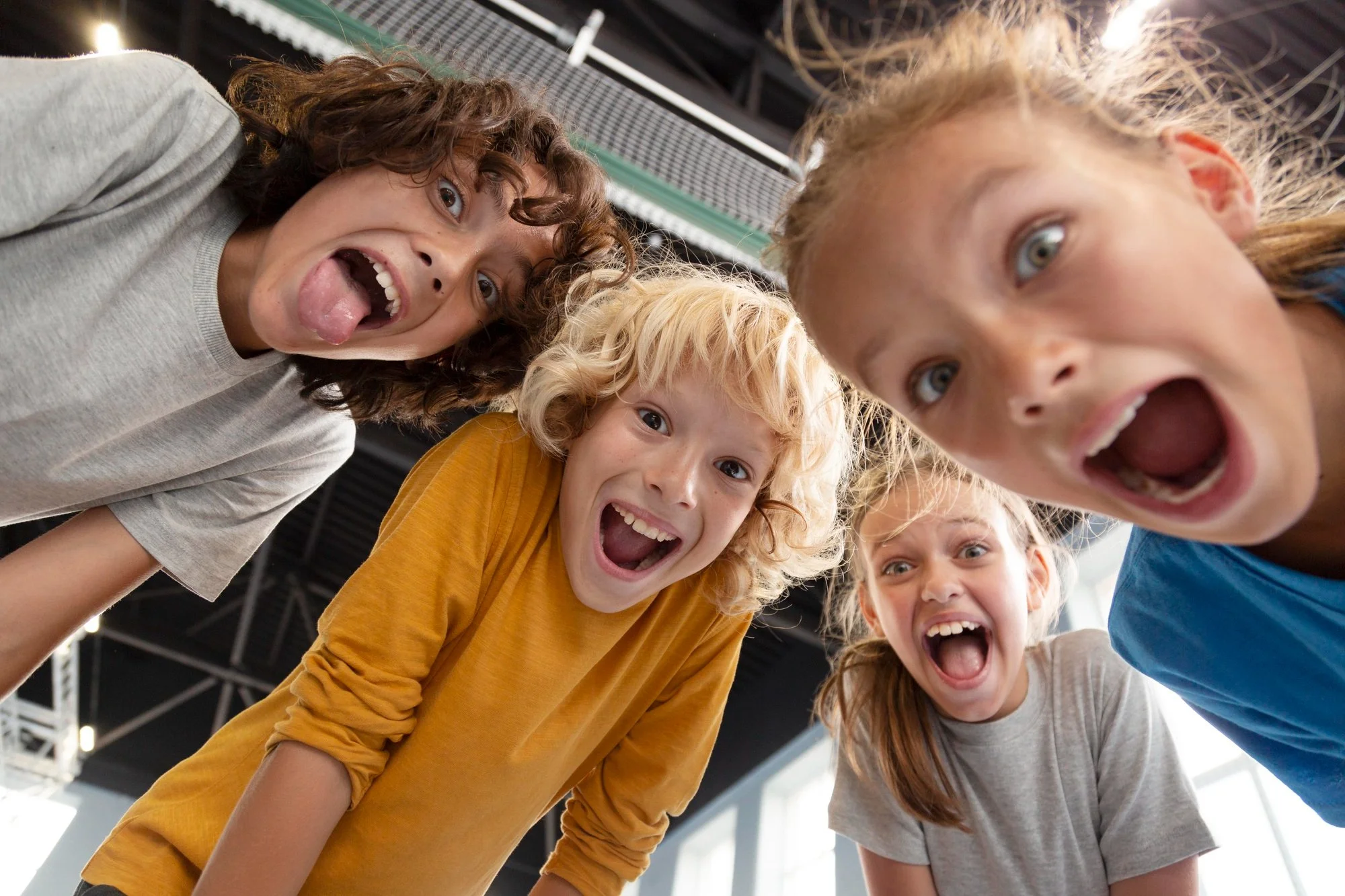 Five children looking down at the camera with surprised and excited expressions. They are in an indoor setting with industrial ceiling lighting.