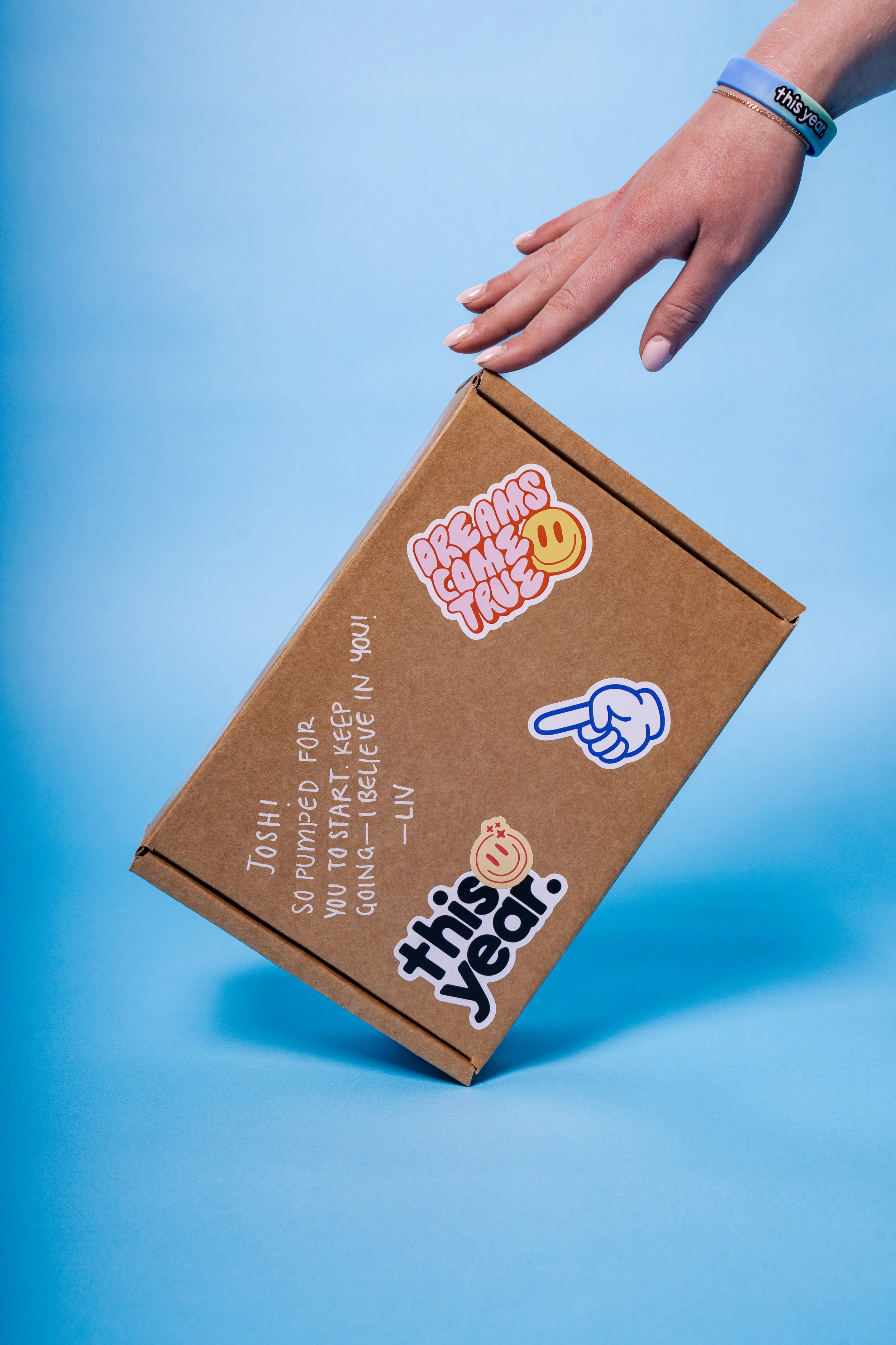 A hand with a blue wristband reaching towards a brown box labeled 'Dreams Come True' with stickers and a handwritten message on a blue background.