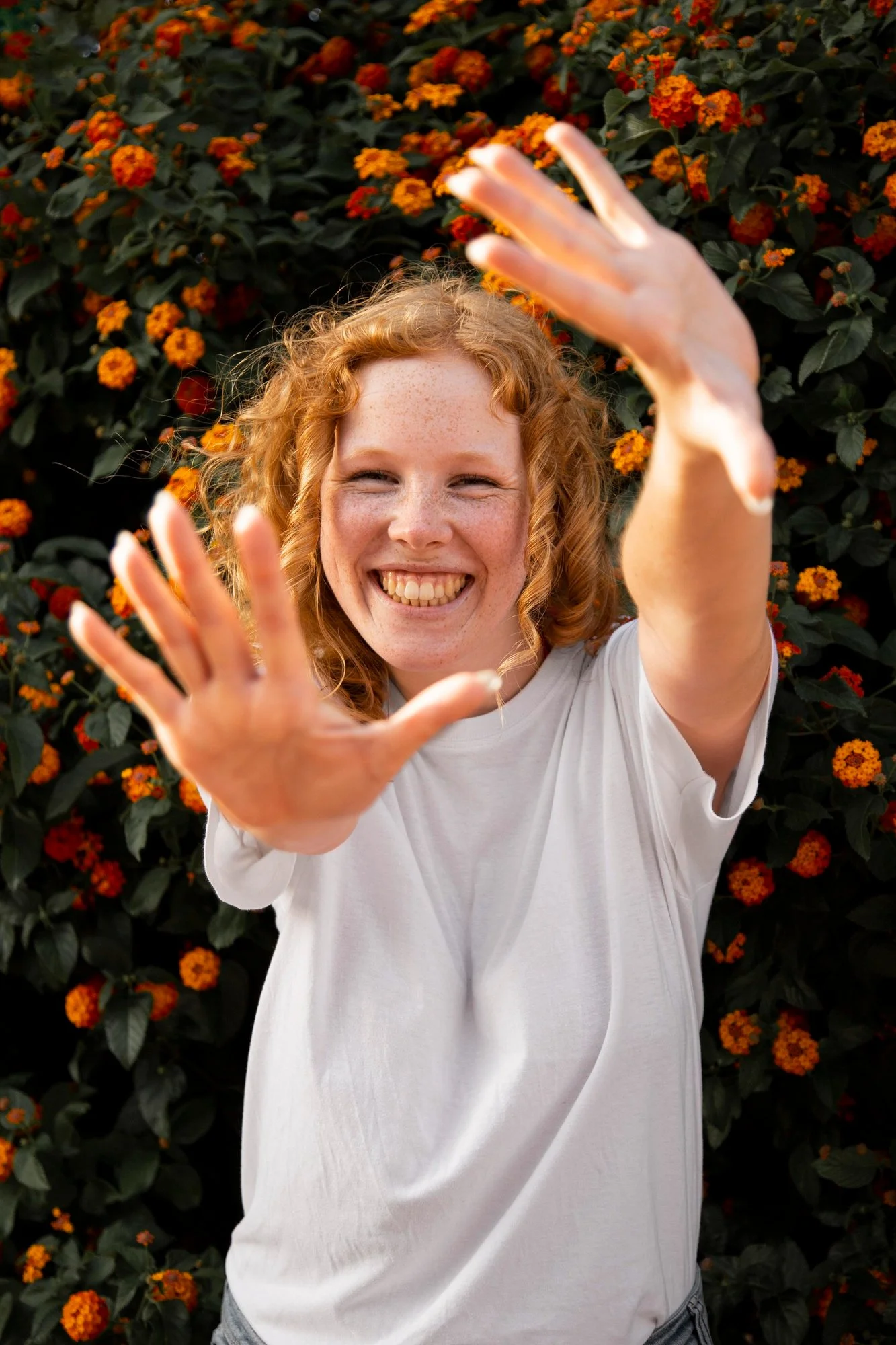 A smiling woman with curly red hair and freckles, extending her hands toward the camera, standing in front of a bush with orange flowers.