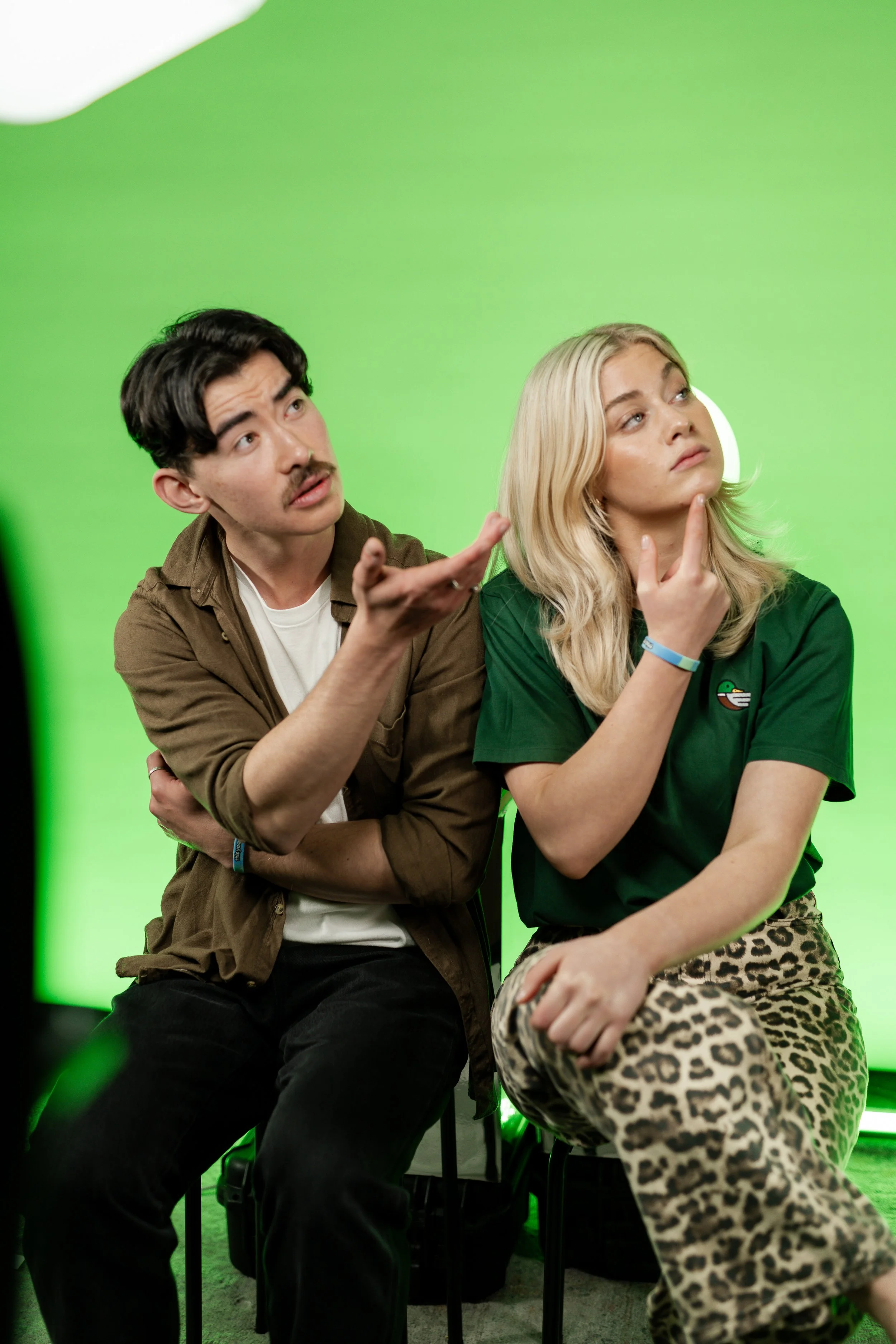 A man and woman sitting on chairs in front of a green screen. The man is gesturing with his hand as he talks, and the woman is sitting thoughtfully with her finger on her chin.