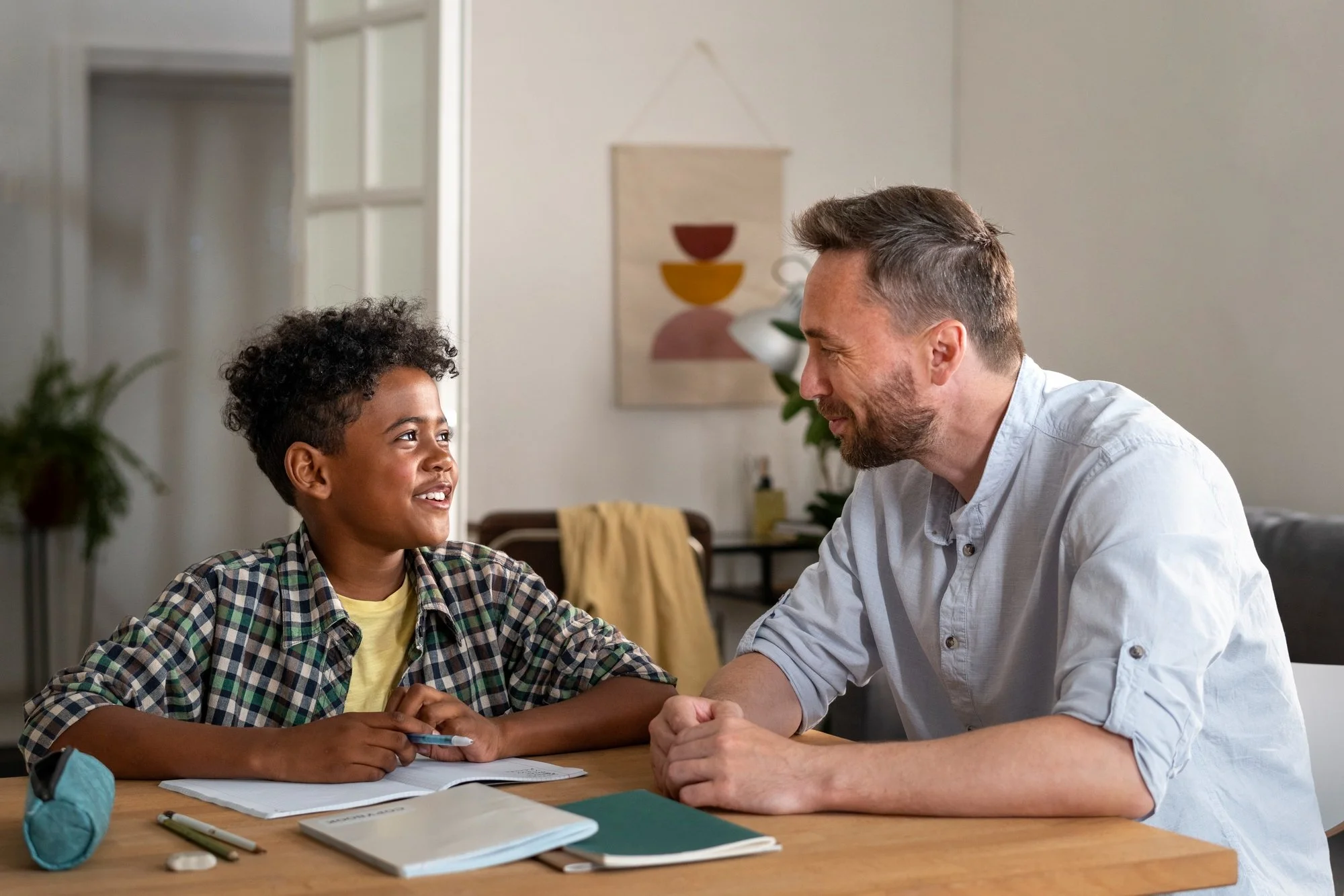 A young boy and an adult man are sitting at a table, smiling and looking at each other, engaging in a conversation in a cozy room with plants and artwork on the wall.