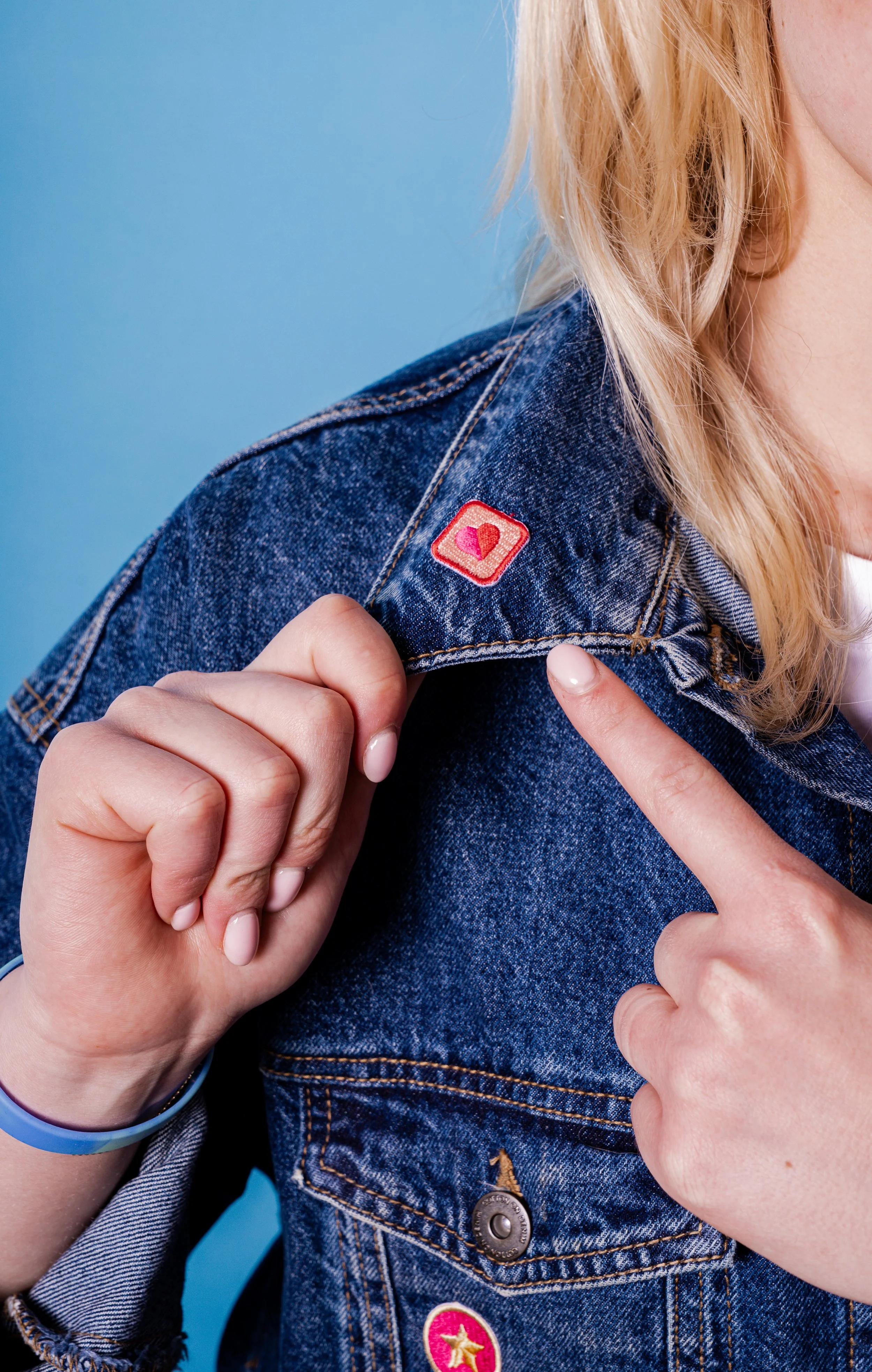 A woman wearing a denim jacket with a pink heart-shaped pin on the shoulder, pointing at her shoulder where the pin is located, against a blue background.