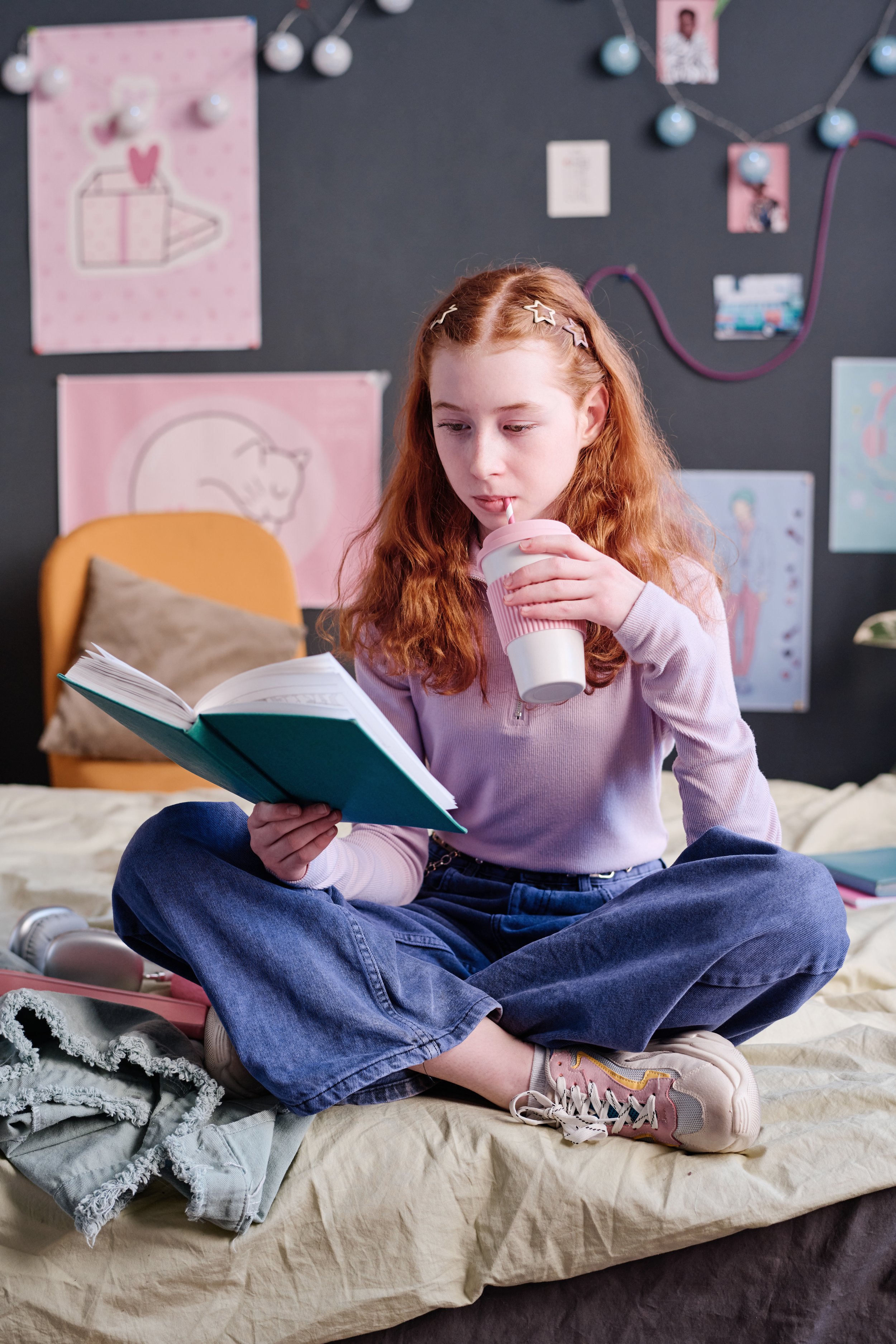 A young girl with red hair and star hair clips sitting on a bed, reading a book and drinking from a pink and white striped cup.