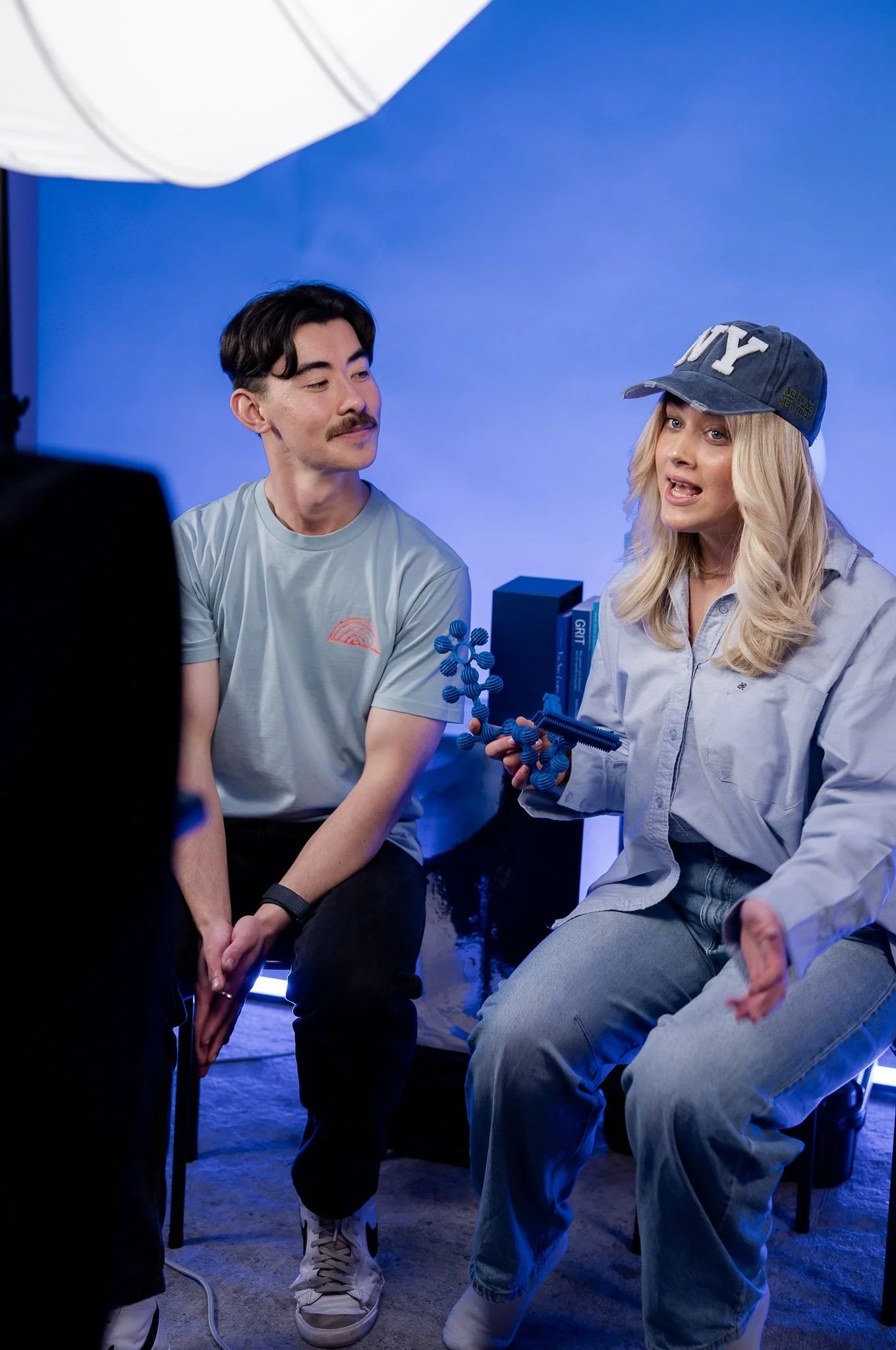 Two people having a conversation indoors, with one woman wearing a blue baseball cap and holding a molecular model, and a man sitting beside her listening attentively.