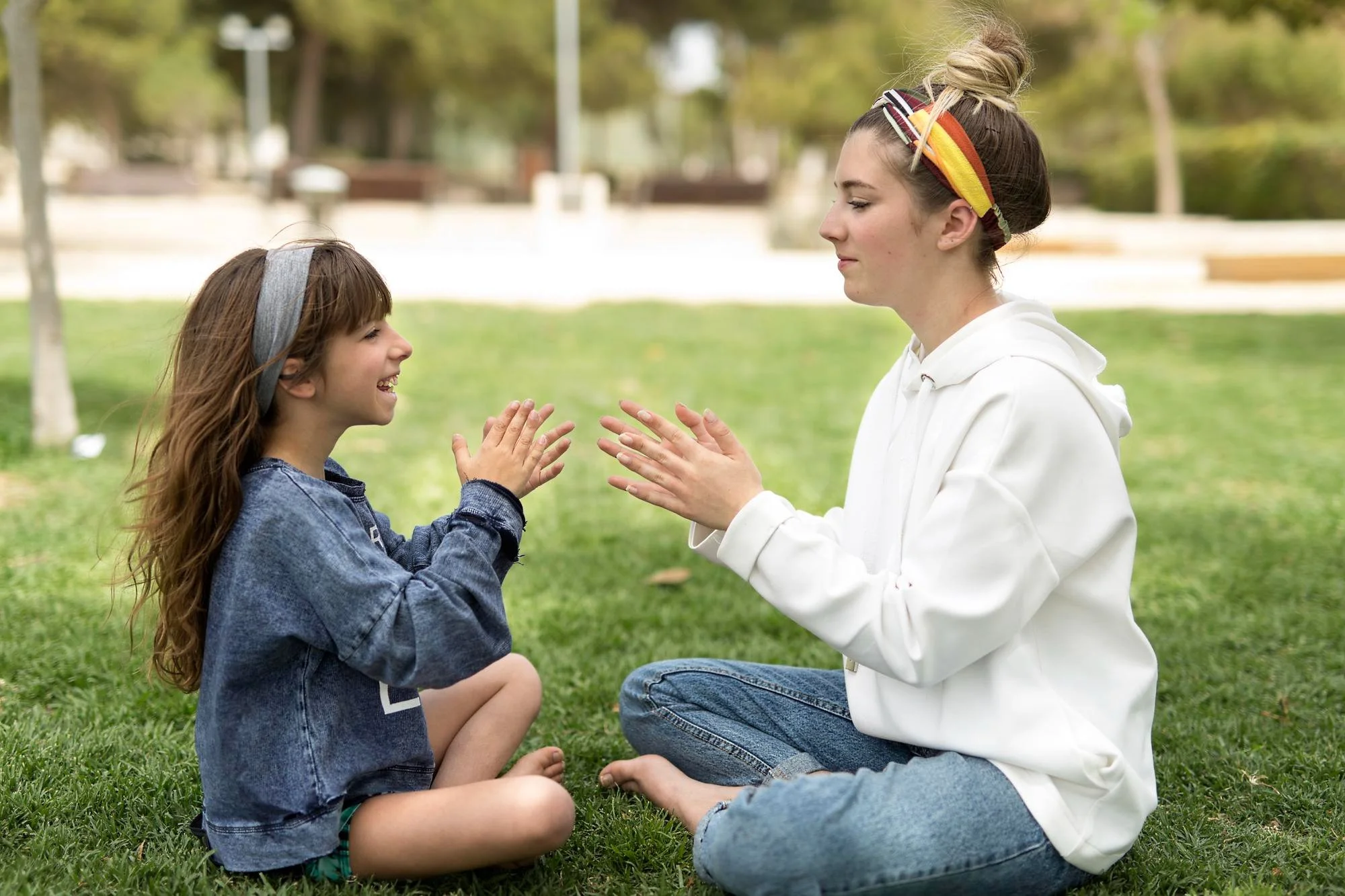 A young girl and woman sit on grass facing each other, clapping and smiling warmly in a park with trees and blurred background.
