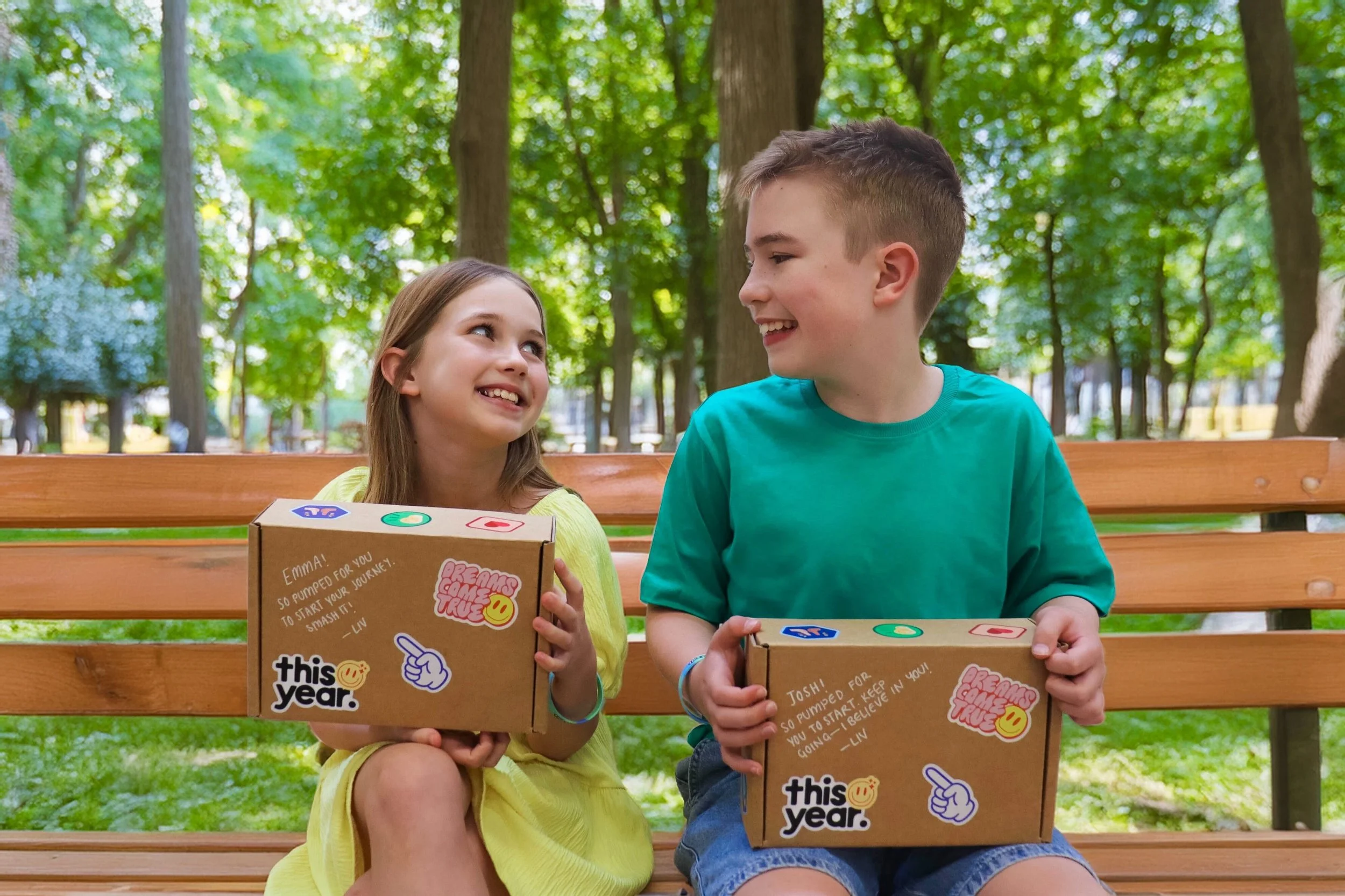 A girl with long brown hair and a boy with short light brown hair sitting on a park bench, smiling at each other, holding gift boxes decorated with colorful stickers and handwritten notes. The background features green trees and a sunny park setting.