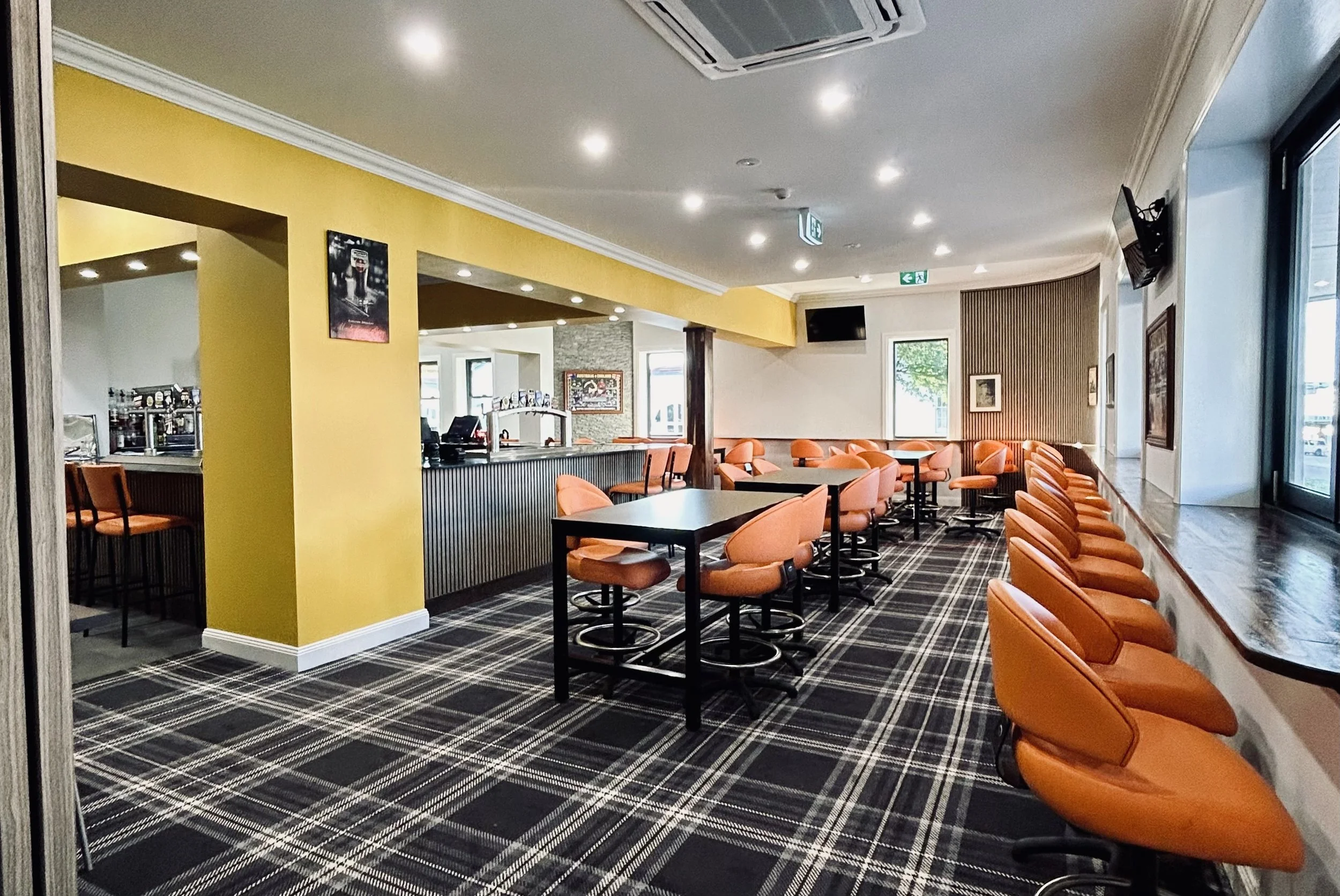 Empty bar and seating area with orange chairs, a yellow accent wall, a black and white checkered carpet, and a bar counter in the background.