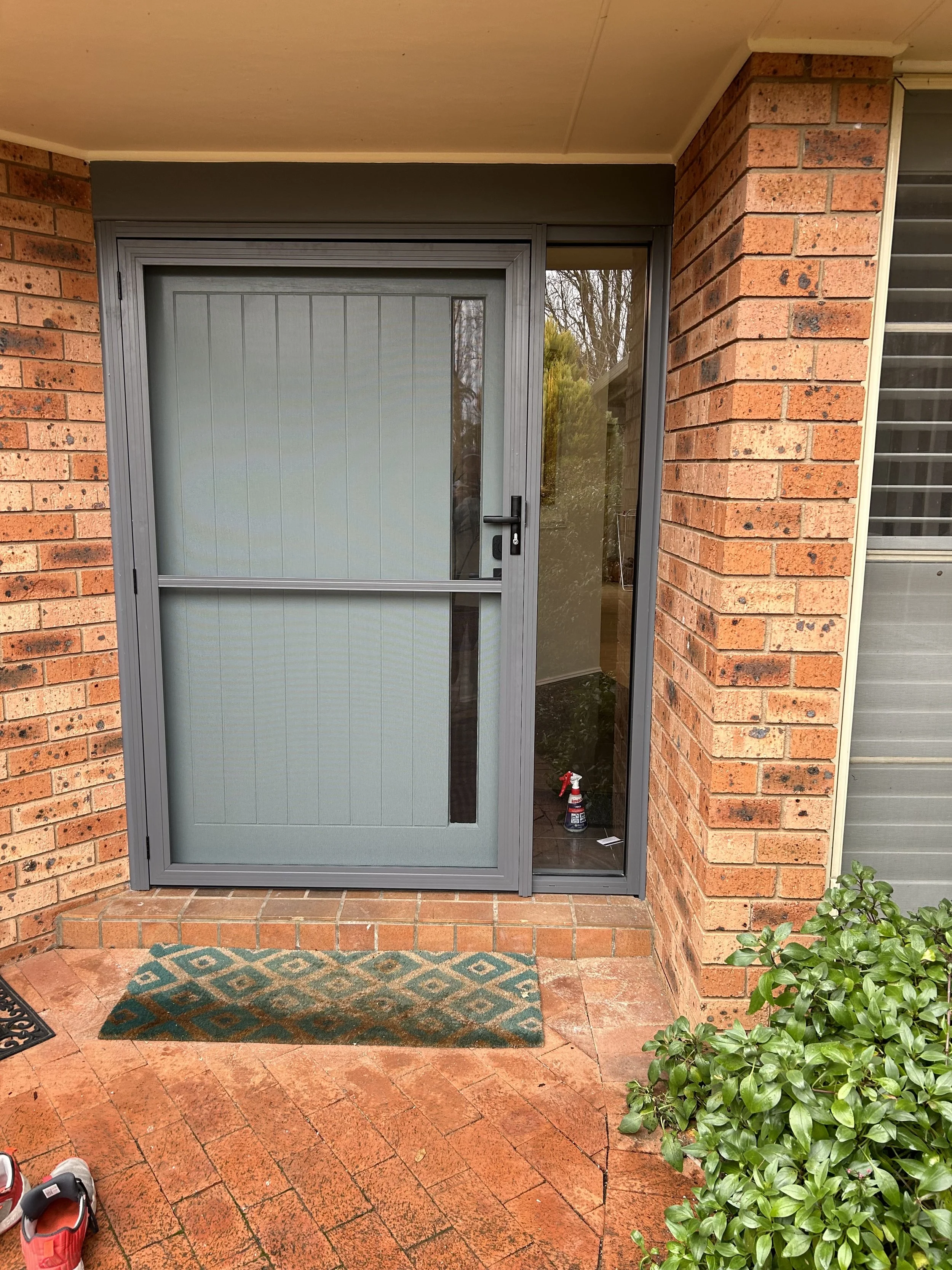 Front porch with brick flooring and walls, a gray screen door with a glass panel and a catch, a small patterned doormat, green bush plants on the right, and a spray bottle on the inside floor near the door.