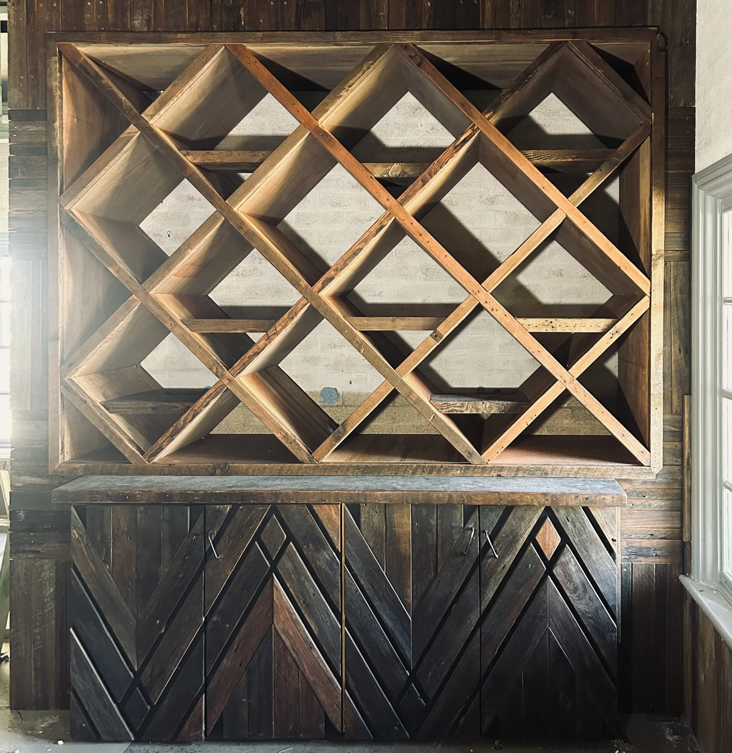Empty wooden wine rack with diagonal and diamond-shaped compartments mounted on a wall, below a wooden storage cabinet with chevron pattern doors.
