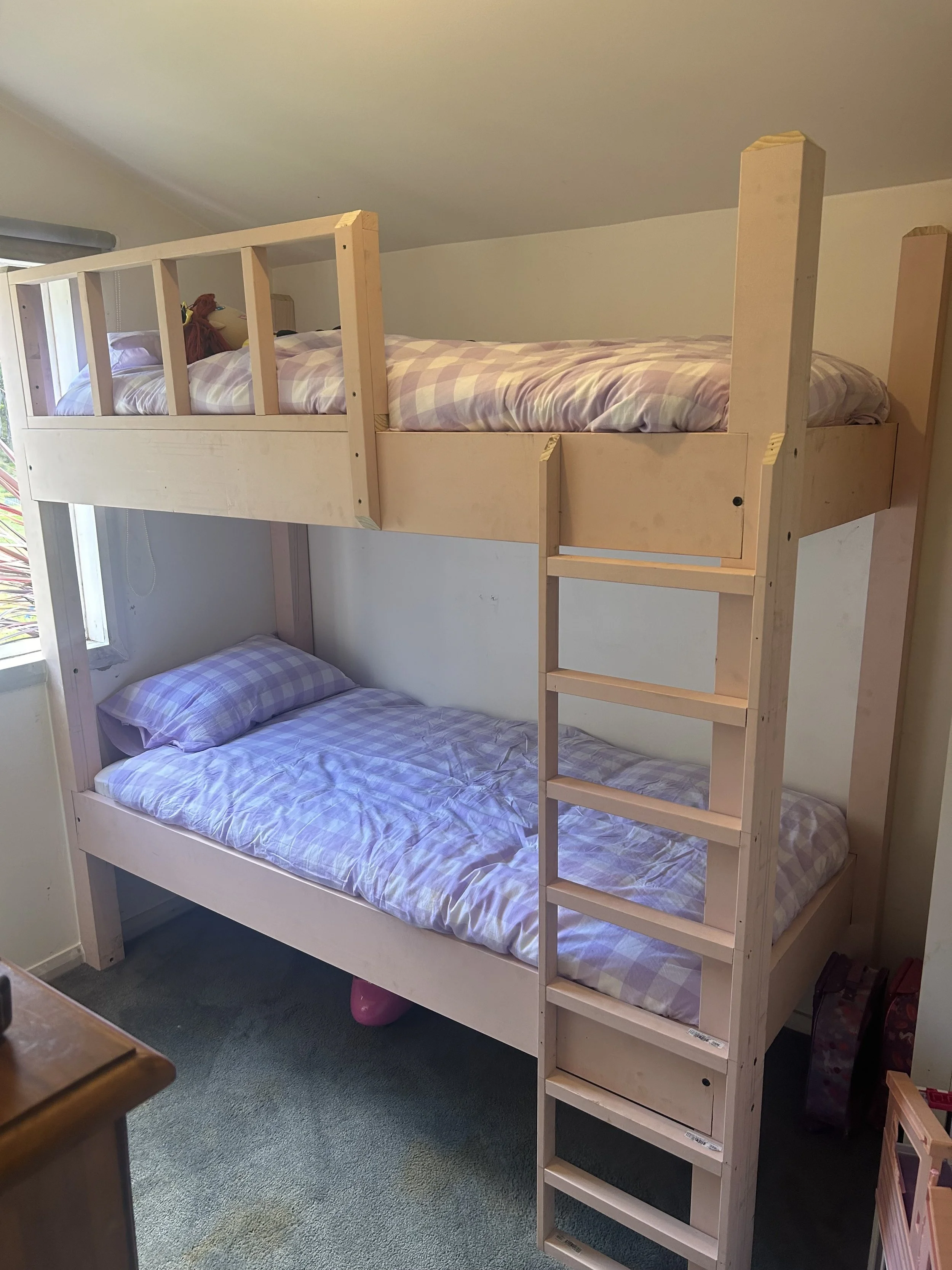A wooden bunk bed with a twin mattress on top and bottom, both with purple checkered bedding, positioned next to a window in a room with carpeted floor.