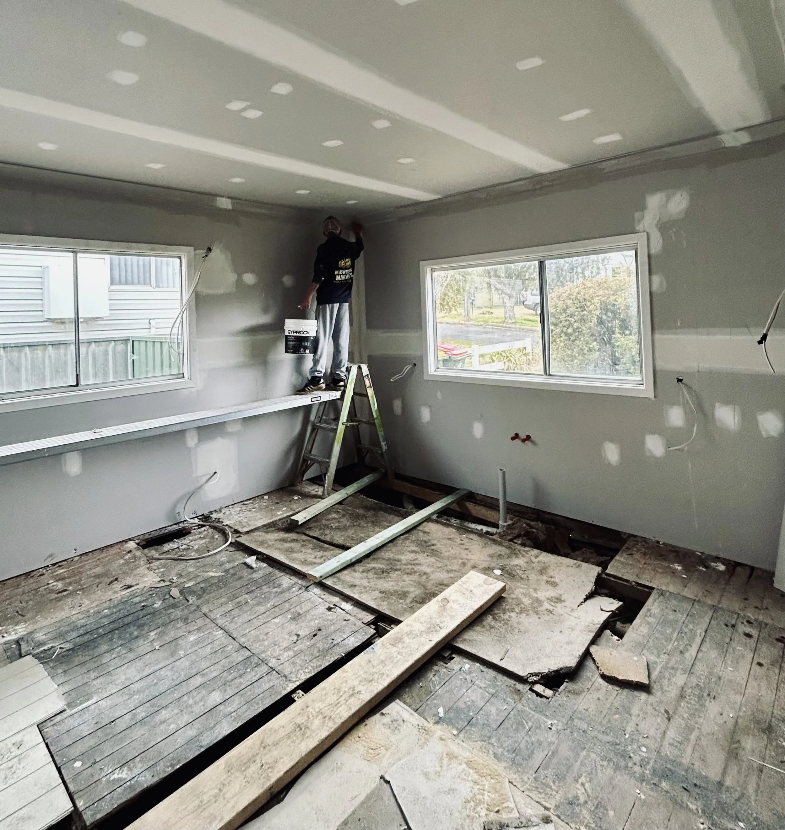 A person working on drywall in a room under renovation, standing on a ladder near the ceiling, with construction tools and exposed flooring visible.
