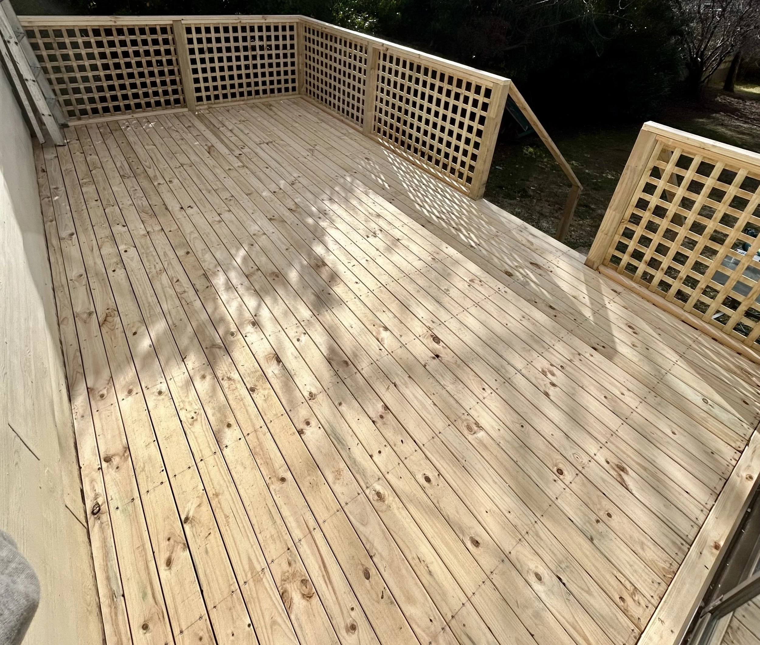 Newly constructed wooden deck with lattice railings, partially in sunlight, adjacent to a house with outdoor trees in the background.