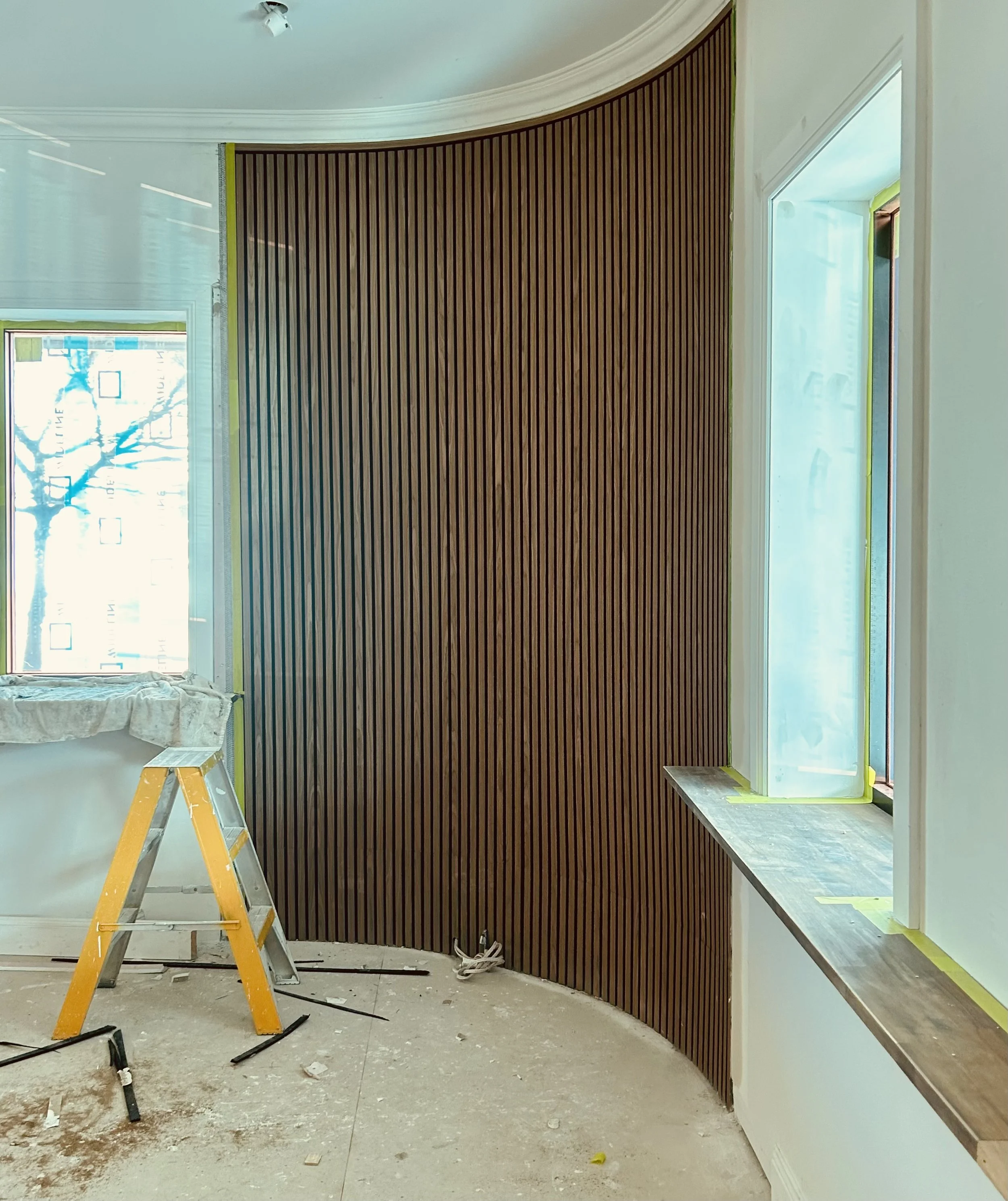 Interior of a room with a curved wooden slat wall, a large window with tape around the frame, and construction tools on the floor.